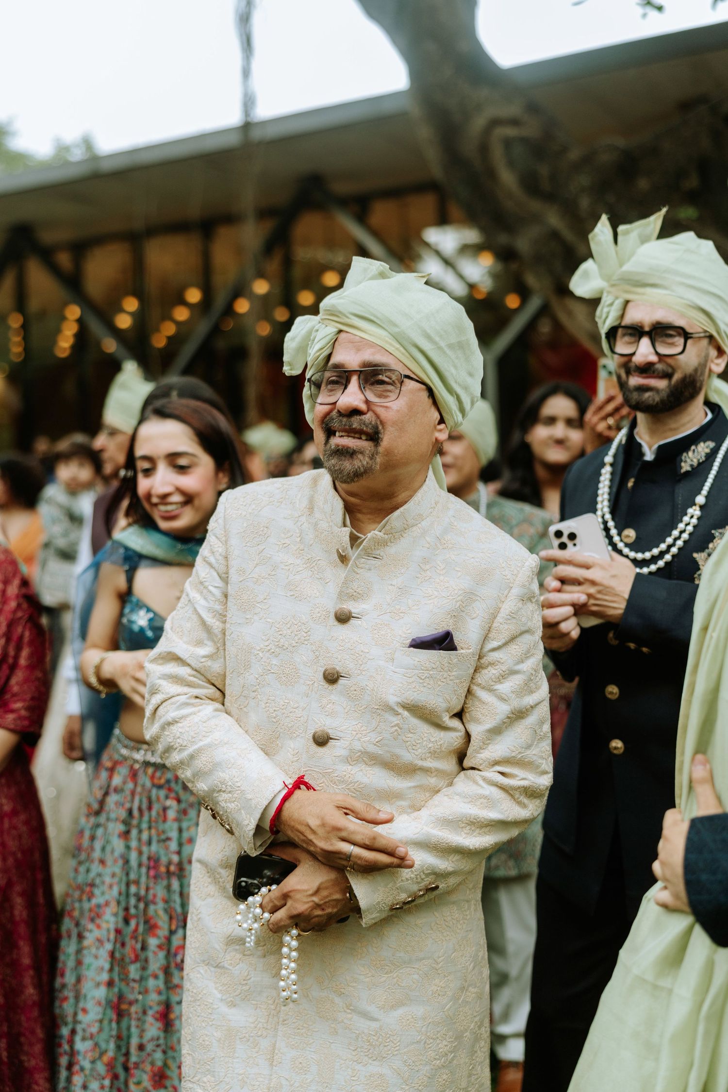 Man in white sherwani and green turban at Indian wedding celebration with guests in traditional attire.