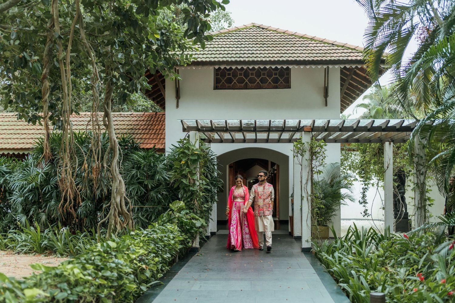 Couple in traditional Indian attire walking through a lush garden pathway at an elegant white villa entrance.