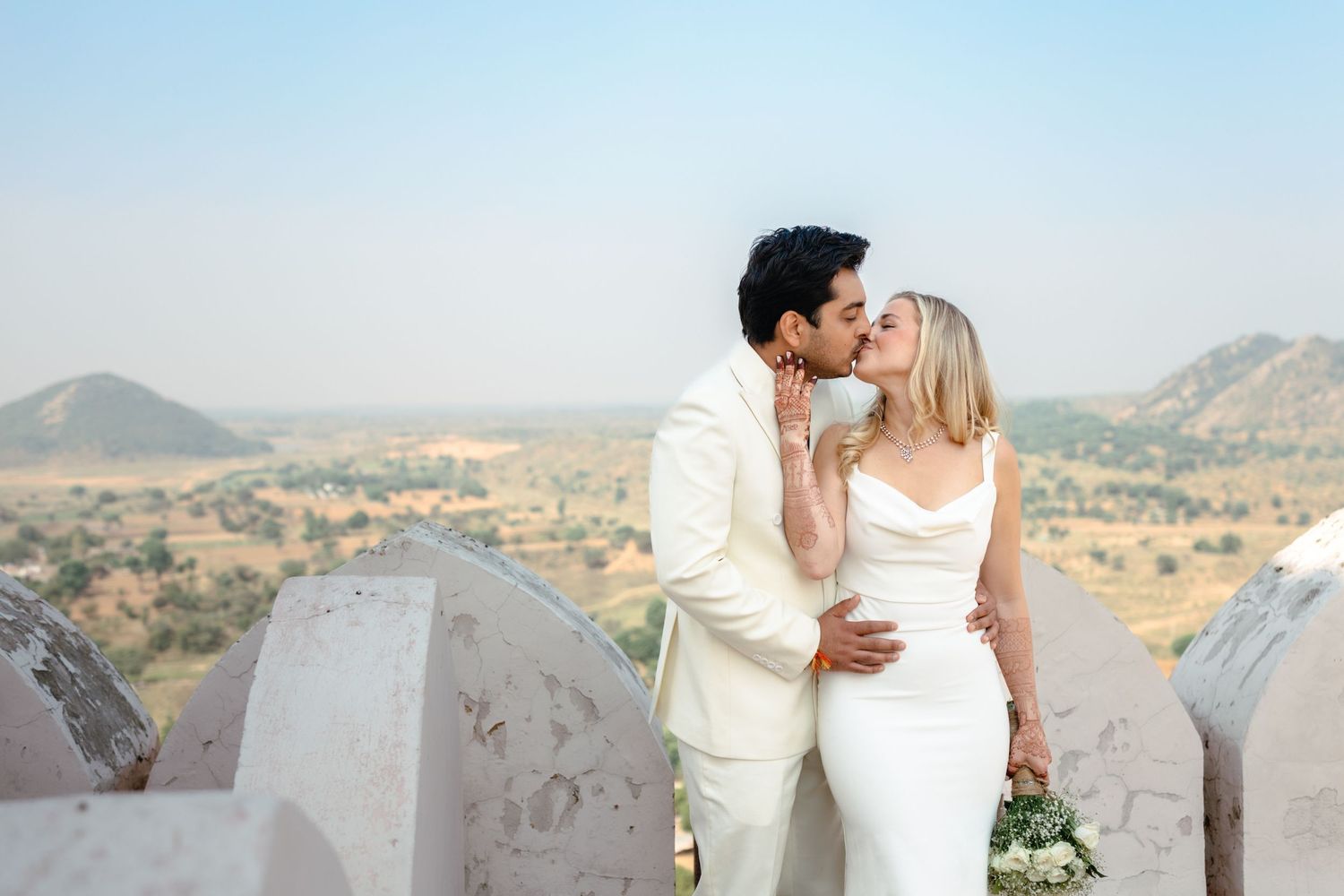 Bride and groom kissing in white wedding attire on a rooftop with scenic desert hills in the background.