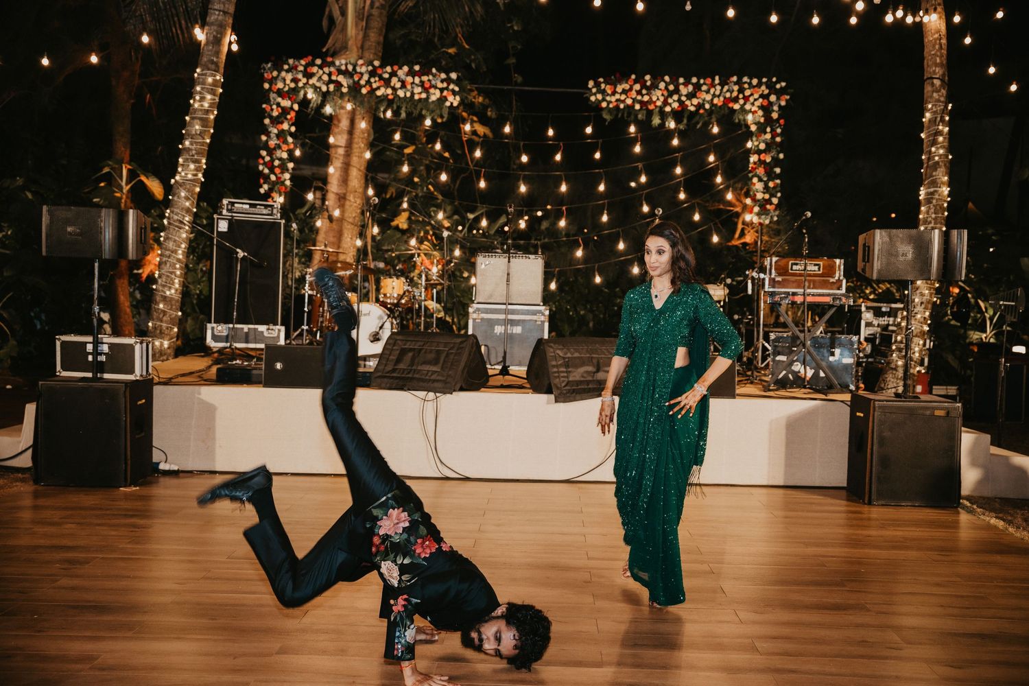 Breakdancer performing headstand on dance floor at elegant outdoor wedding reception with string lights.