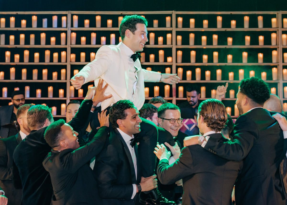 Wedding guests raise their glasses in celebration against a dark backdrop with wooden shelving.