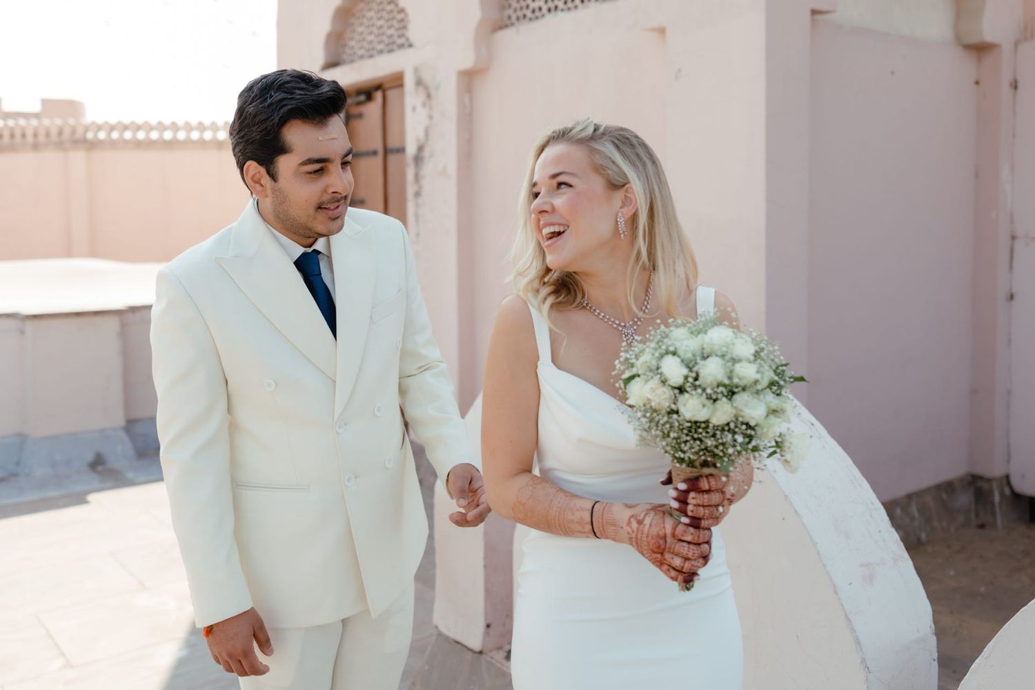 Newlyweds share a joyful moment against pink walls with white floral bouquet.
