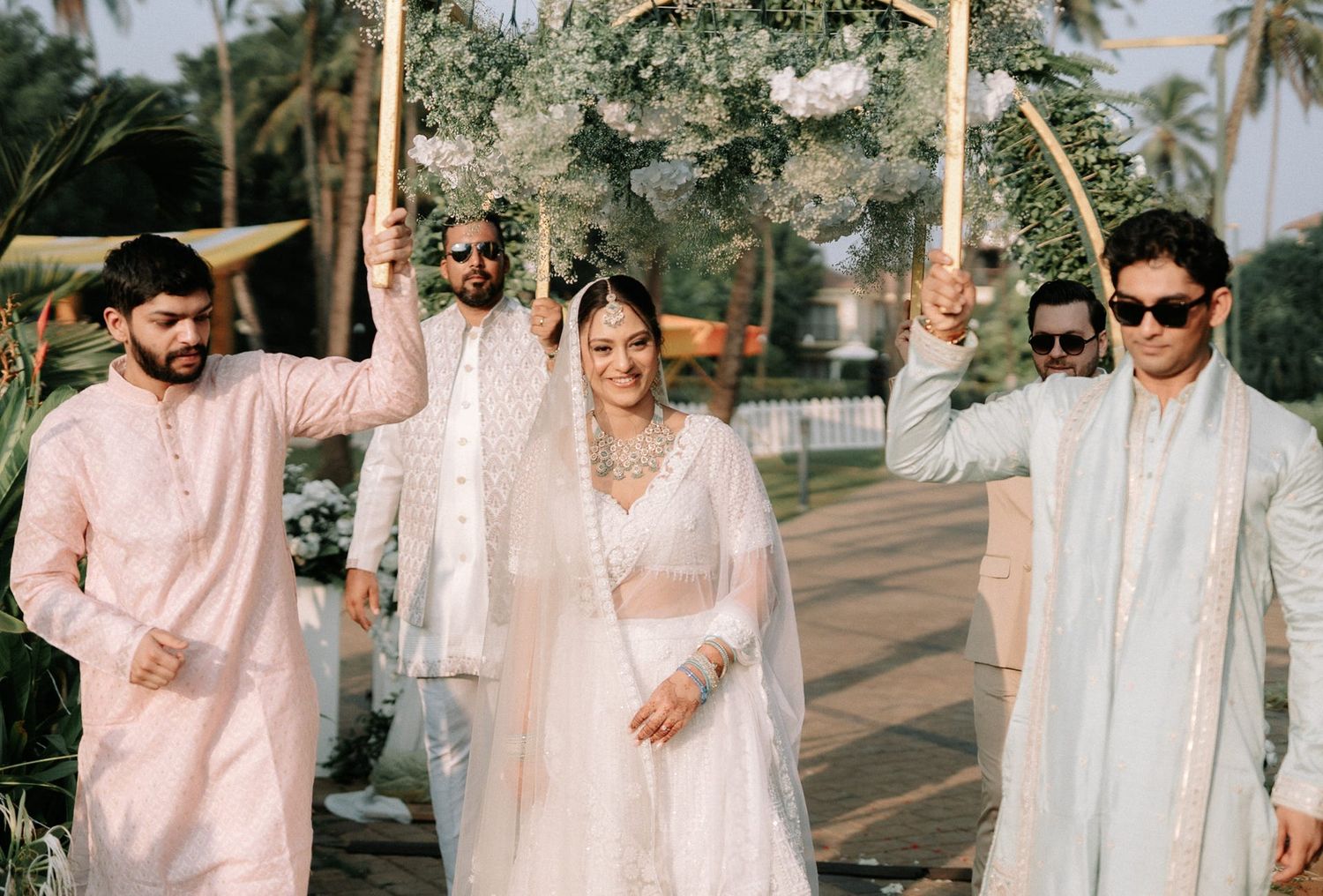 Bride in white lehenga walks with groomsmen holding floral canopy at elegant Indian wedding ceremony outdoors.