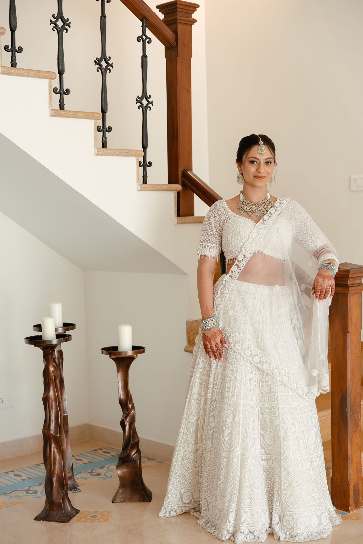 Bride in white embroidered lehenga with dupatta and silver jewelry poses by staircase with candle holders.