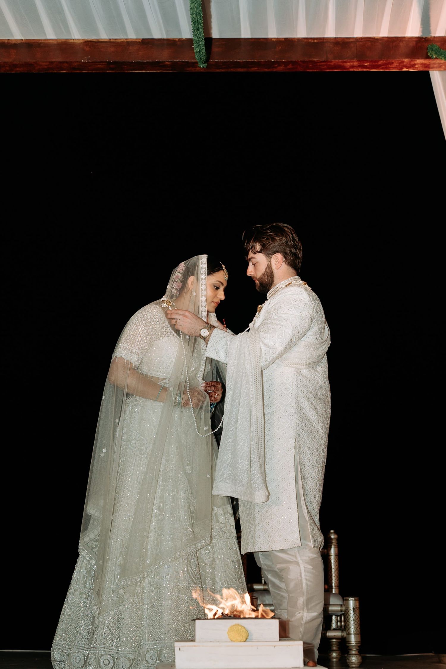 Couple in white traditional Indian wedding attire stand facing each other near a sacred fire during a Hindu ceremony.
