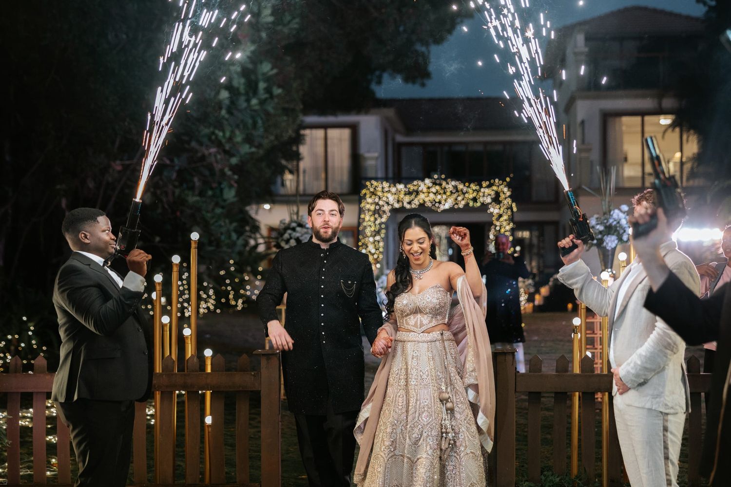 Couple walks through sparkling cold fireworks display at romantic outdoor Indian wedding celebration.