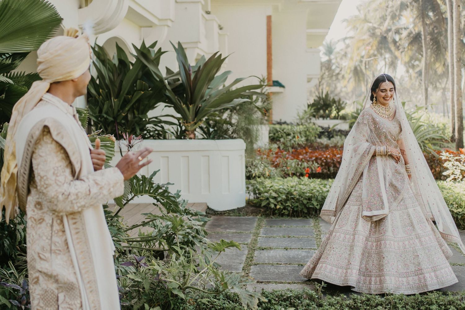 Two people in elegant traditional Indian wedding attire stand in a lush garden setting with white and cream decor.