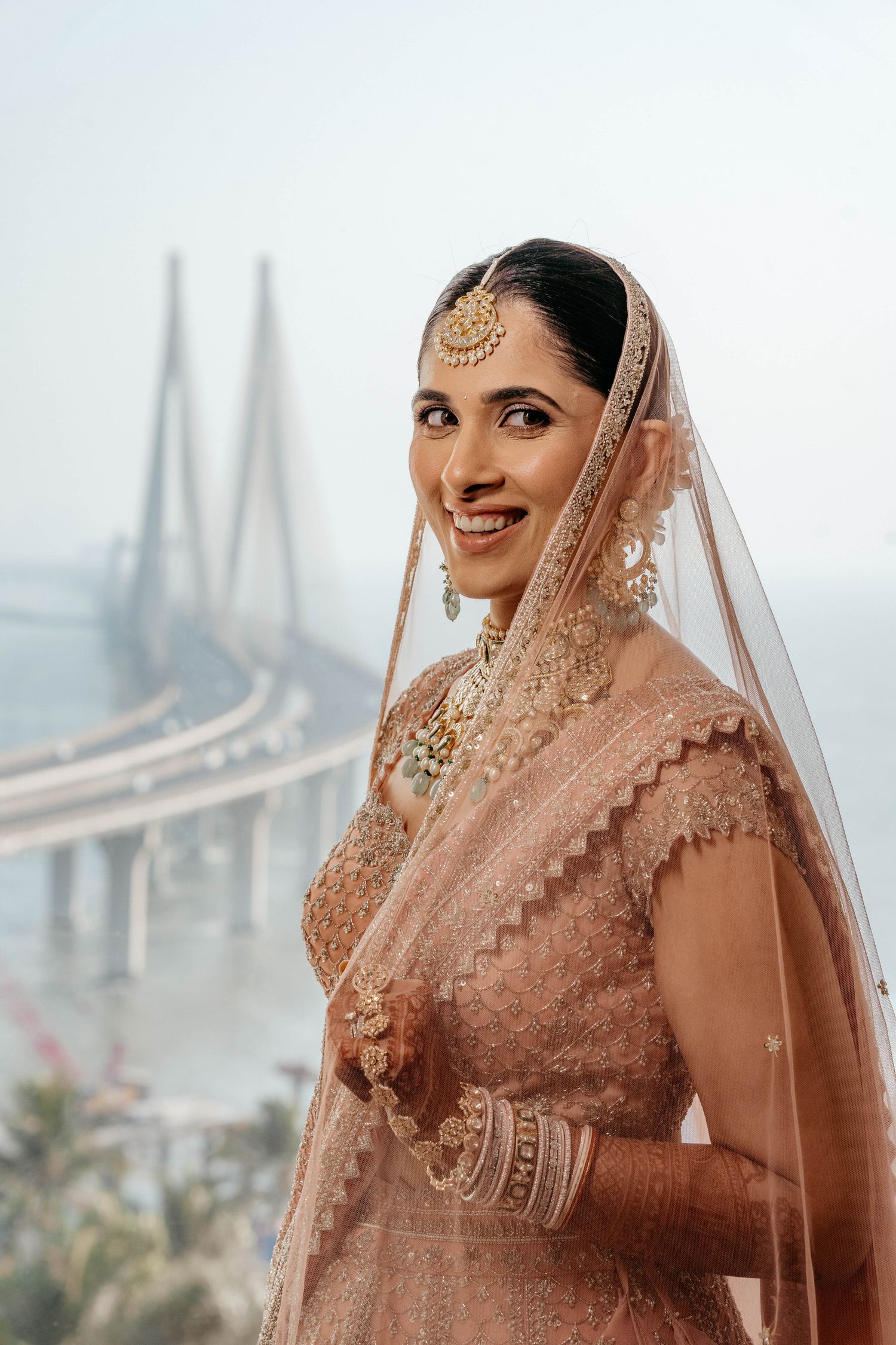 Traditional Indian bride in peach lehenga poses against Mumbai's iconic Bandra-Worli Sea Link bridge backdrop.