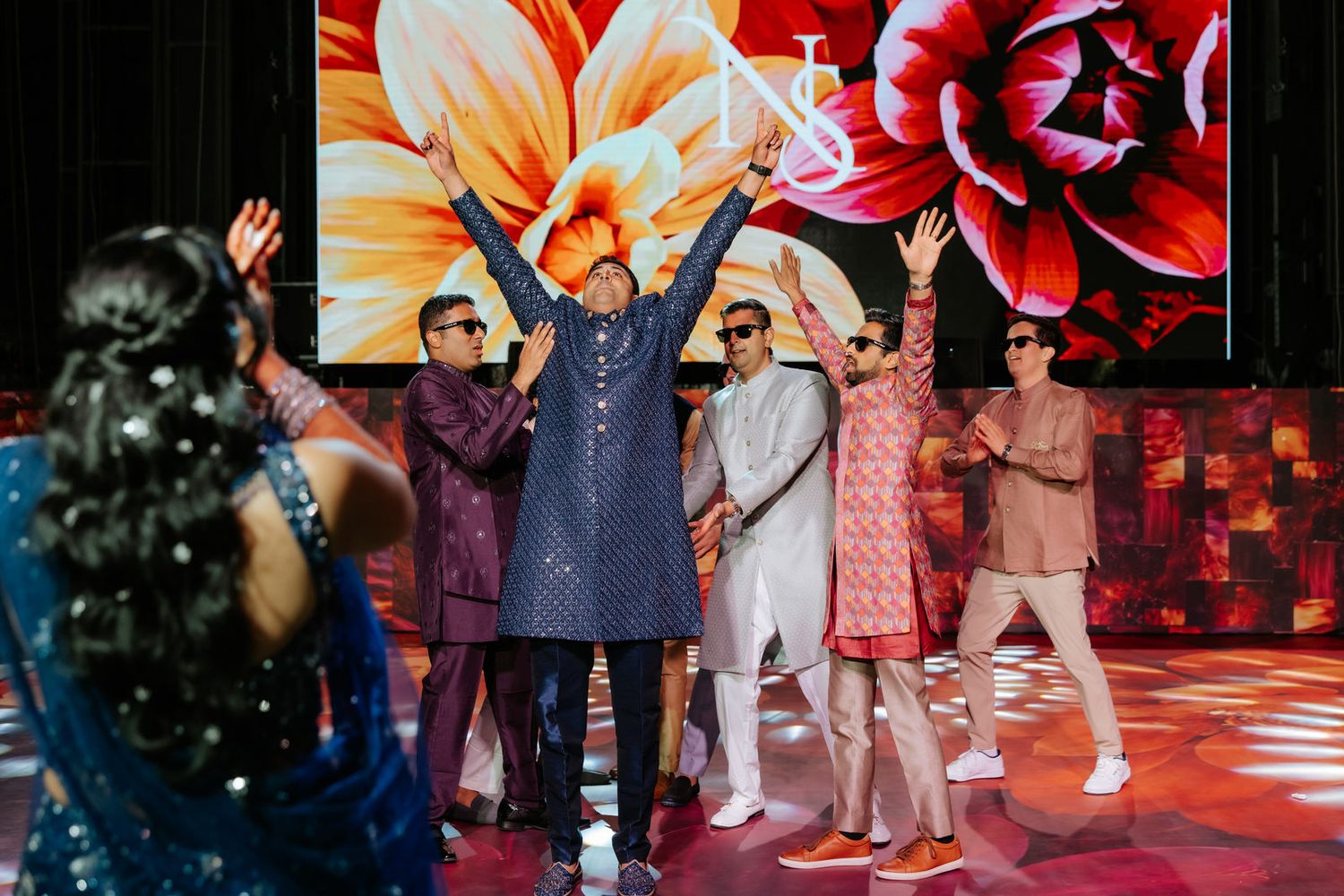 Men in colorful traditional Indian kurtas celebrate on stage with raised arms at a vibrant wedding reception.