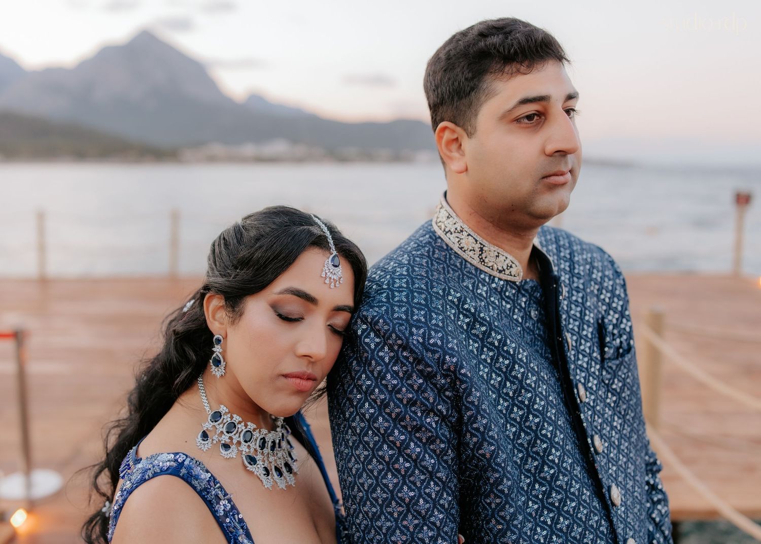 Couple in matching blue Indian attire posing by a scenic waterfront with mountains in the background.