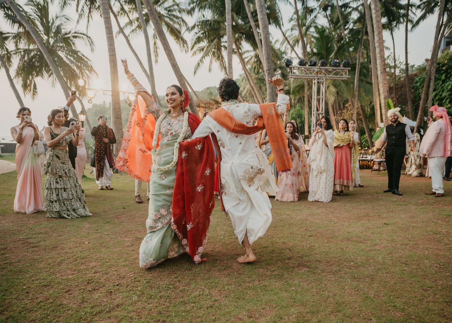 Women in colorful sarees dancing joyfully at an outdoor Indian wedding celebration surrounded by palm trees.