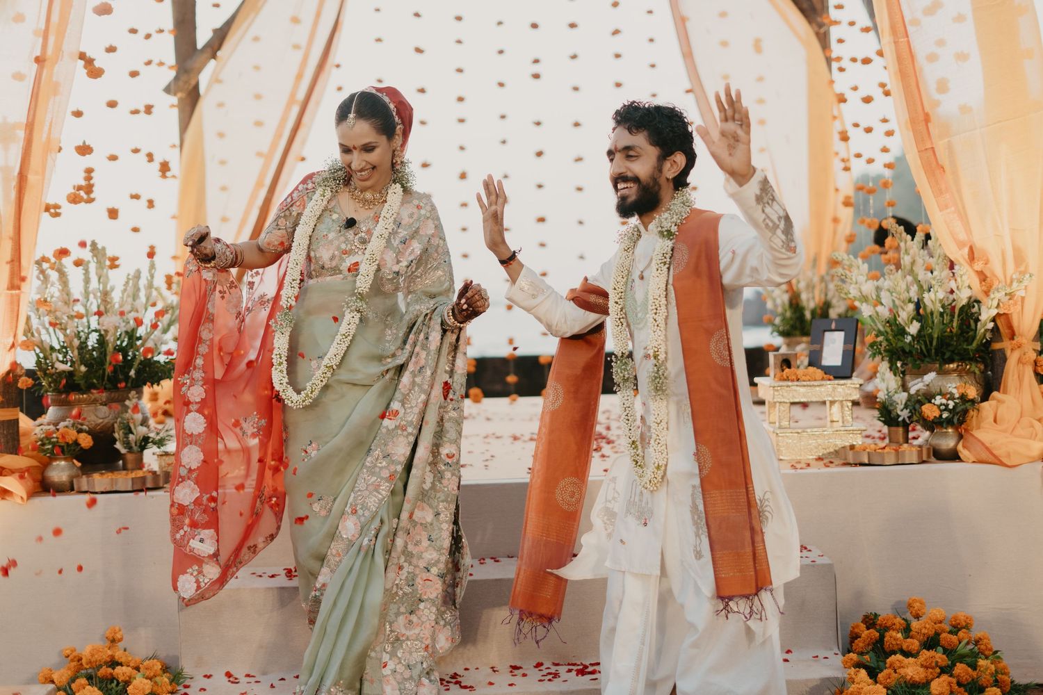 Joyful couple in traditional Indian wedding attire celebrating under a floral mandap with marigold decorations.