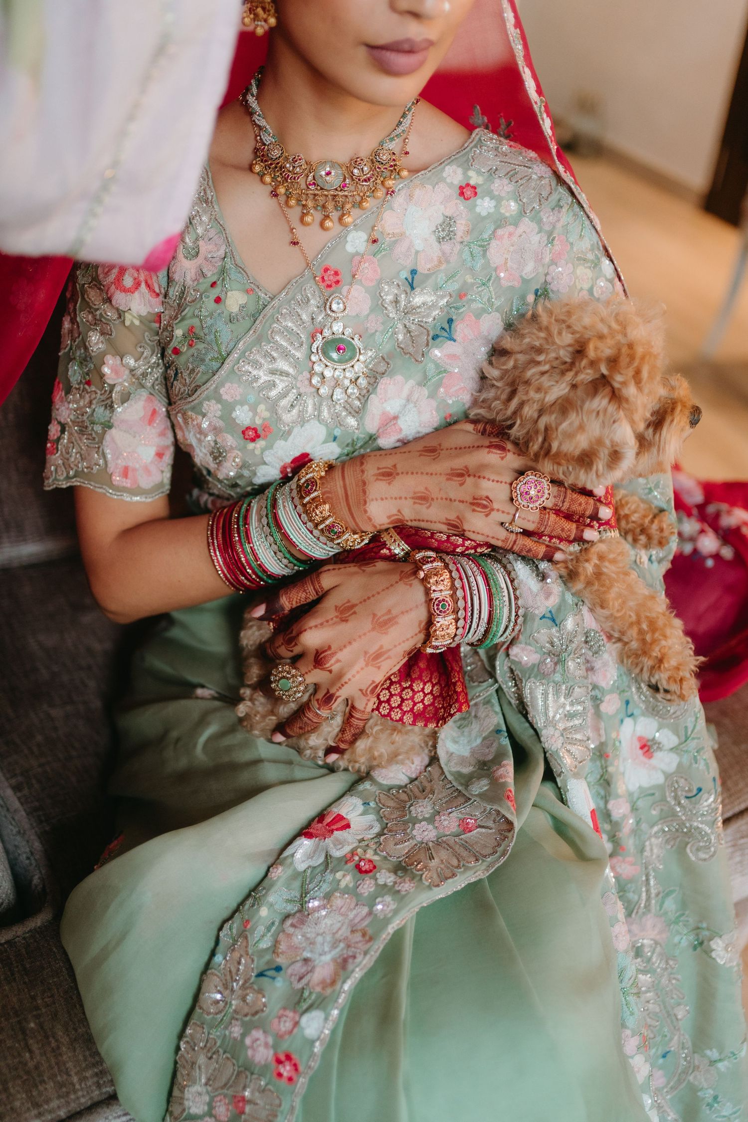 Bride in mint green floral saree with gold jewelry, mehndi hands, and bangles holding a small fluffy dog.