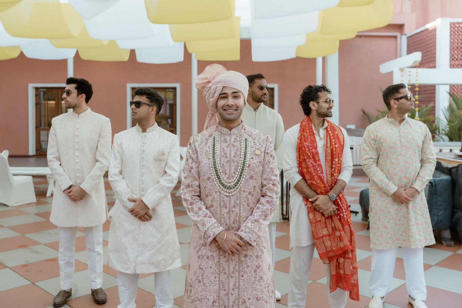 A group wearing traditional Indian wedding attire poses together in a line against a pink wall.