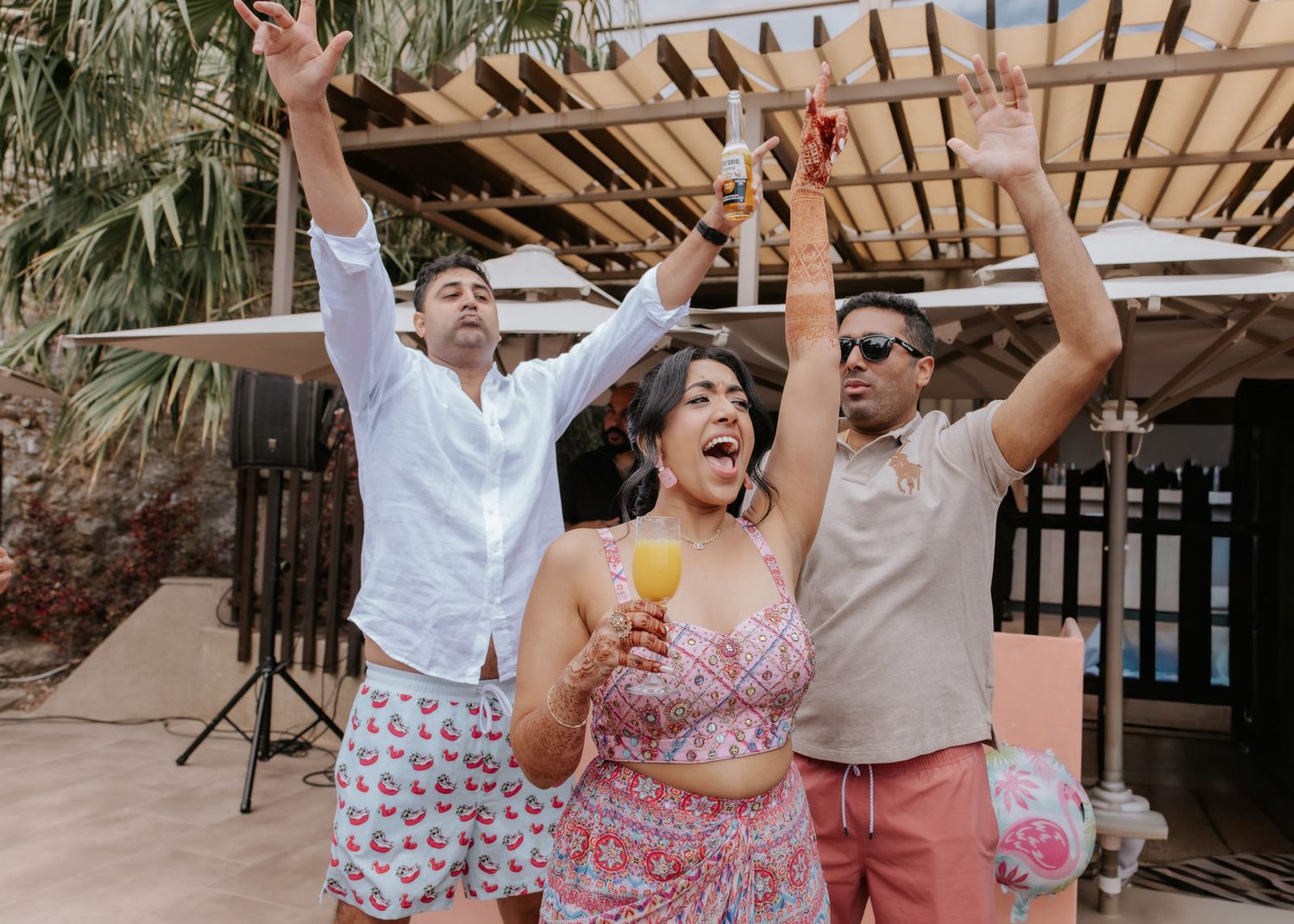 Three friends celebrate outdoors, raising hands and holding drinks at a tropical party venue.
