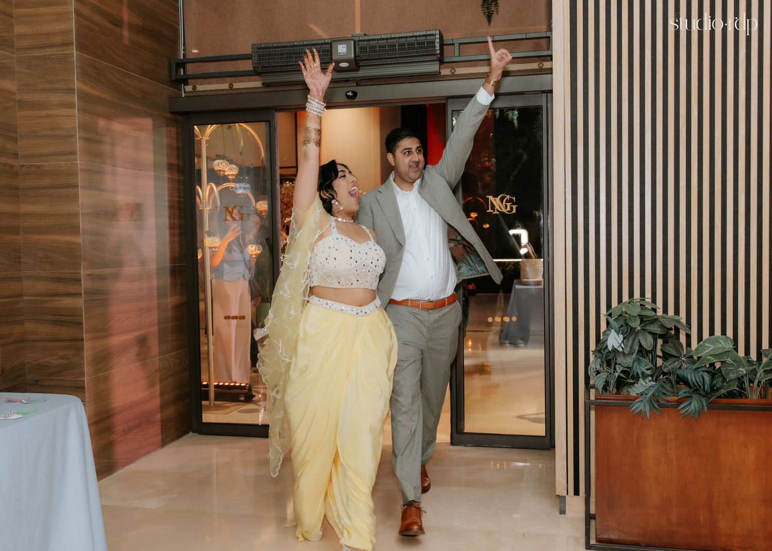 Couple in festive Indian attire waving joyfully at their wedding reception entrance.