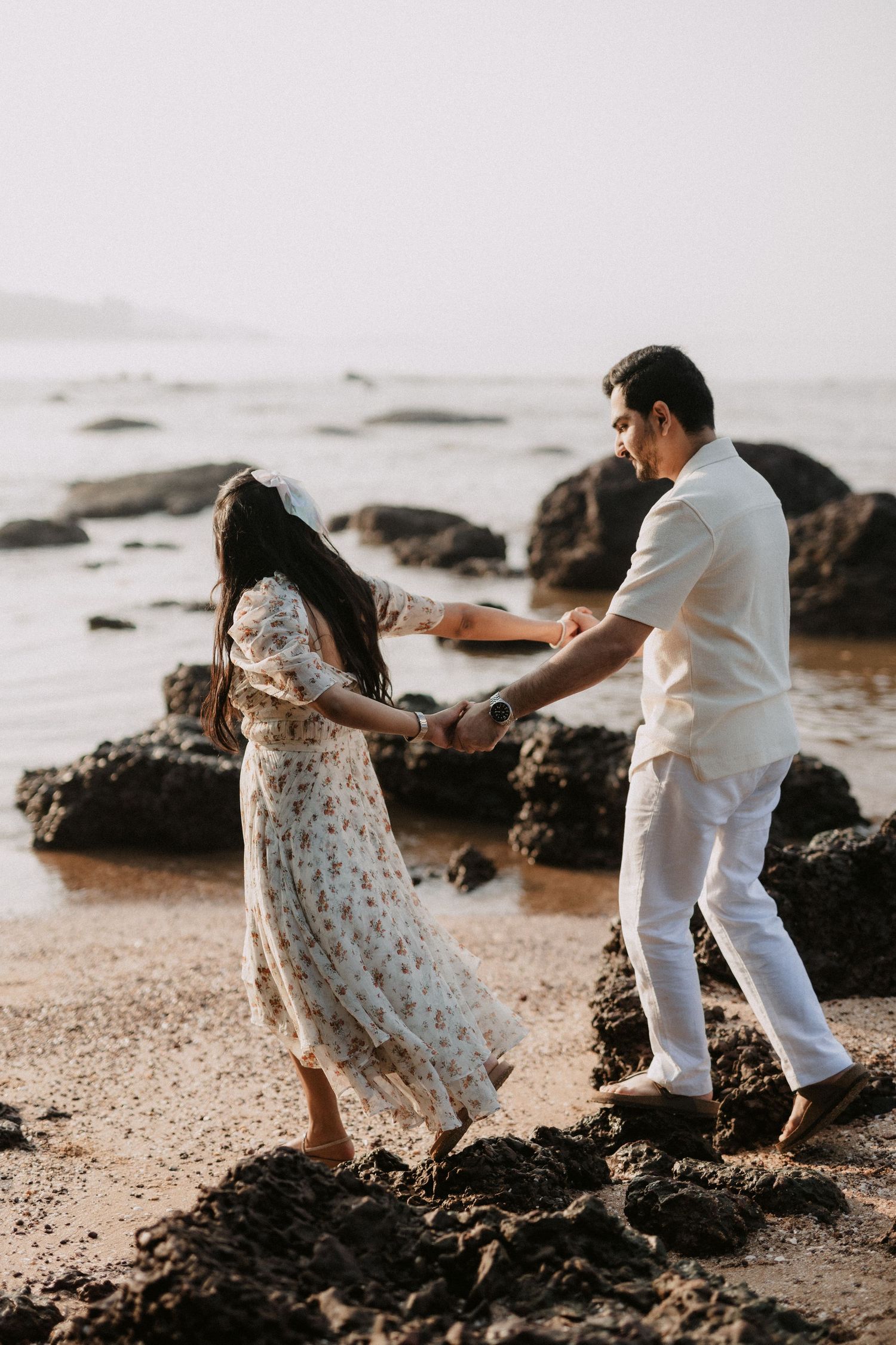 A couple plays and dances together on a rocky beach at sunset along the ocean's edge.