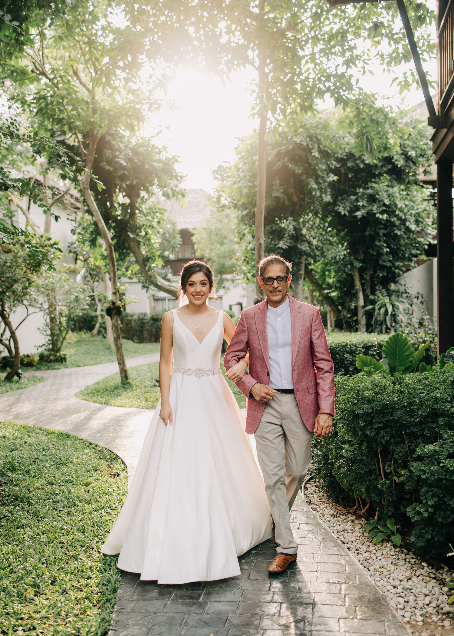 A couple in wedding attire walks hand in hand along a stone pathway surrounded by lush greenery on a sunny afternoon.