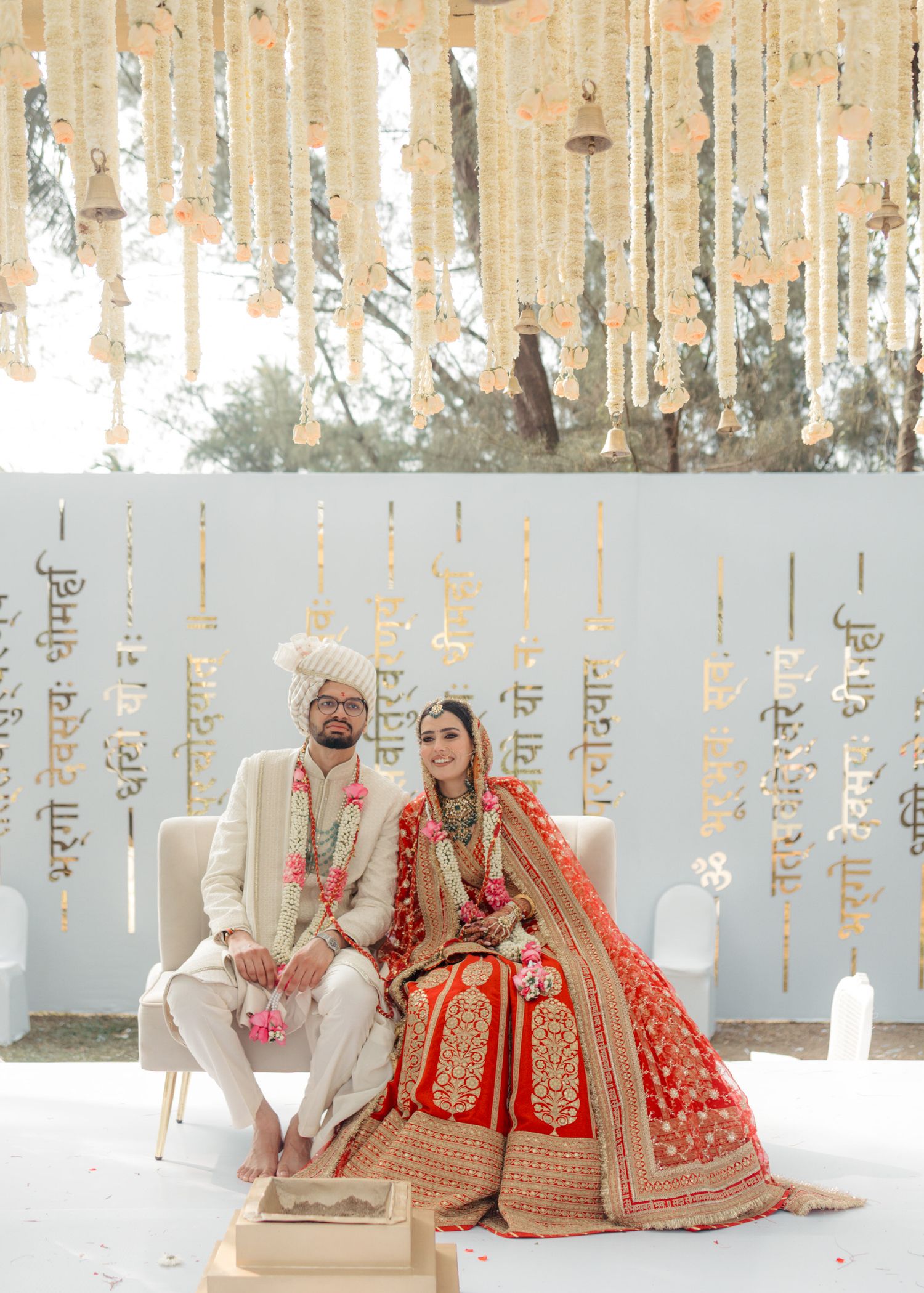 Couple in traditional Indian wedding attire sits against a white and gold decorated backdrop at their ceremony.