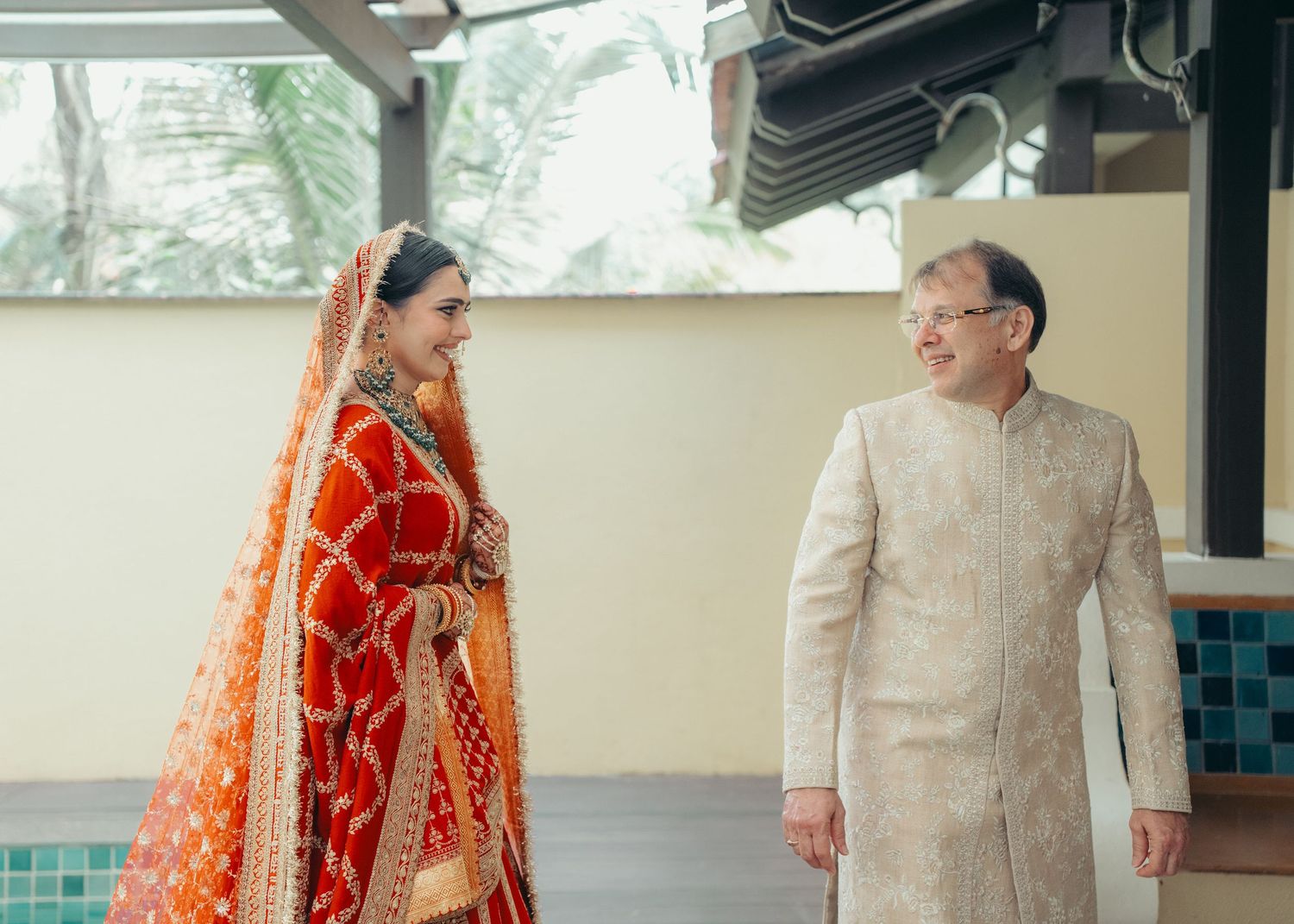 Couple in traditional Indian wedding attire walking together down a covered walkway outdoors.