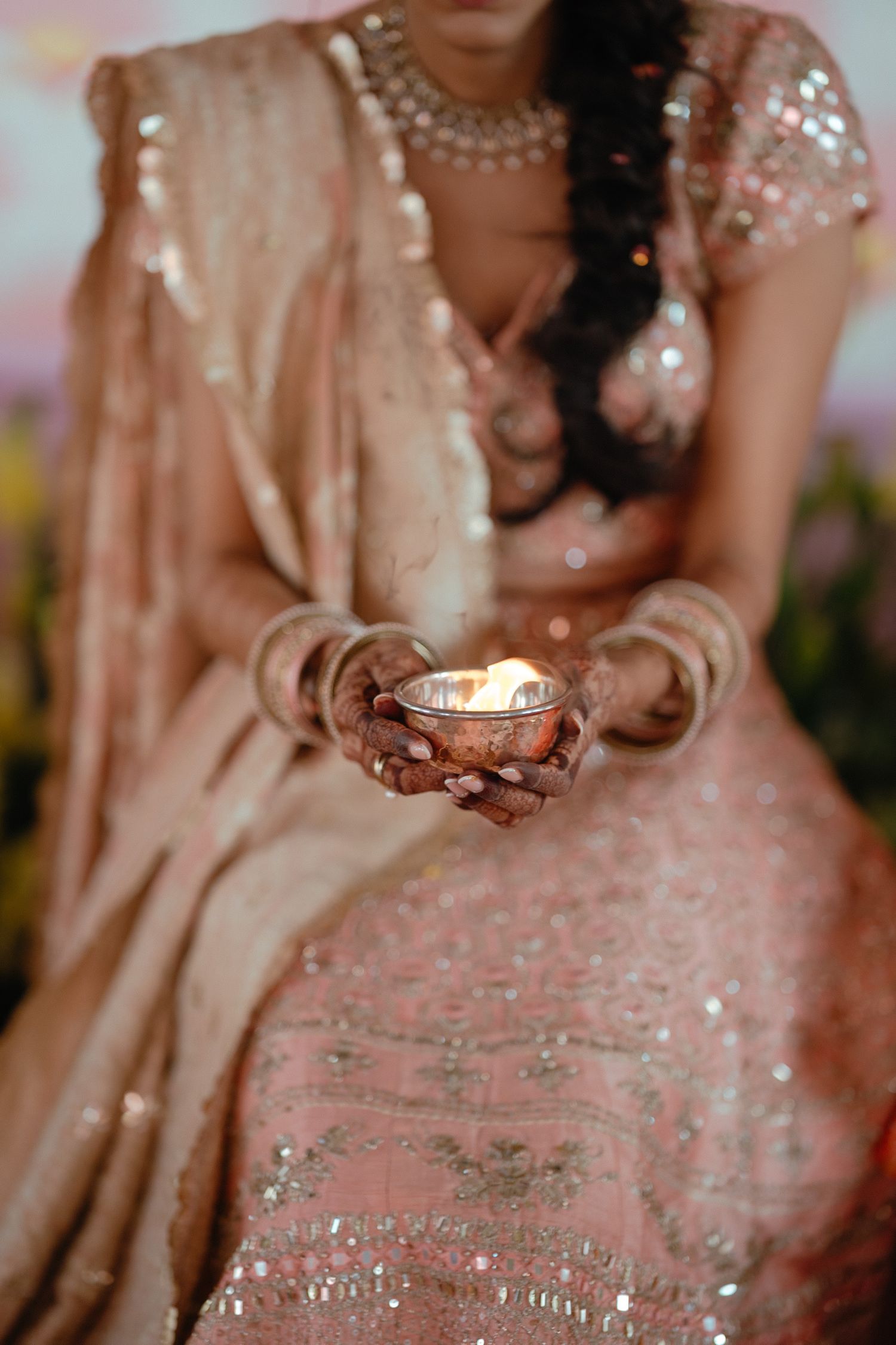 Hands hold a lit diya lamp while wearing a shimmering pink sequined lehenga and pearl jewelry for Diwali celebration.