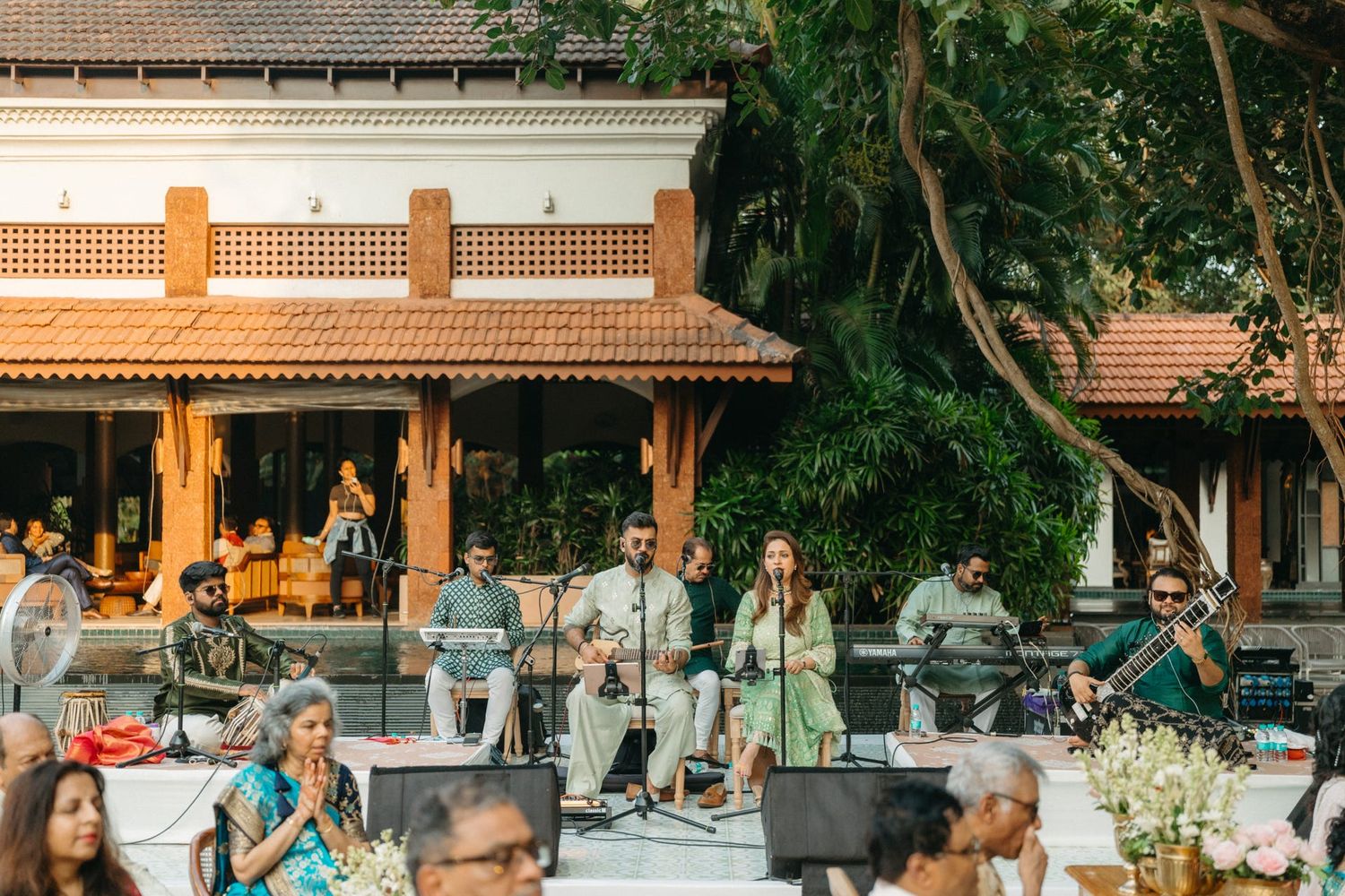 Live band performing at an elegant outdoor Indian wedding reception surrounded by lush trees and a traditional venue.