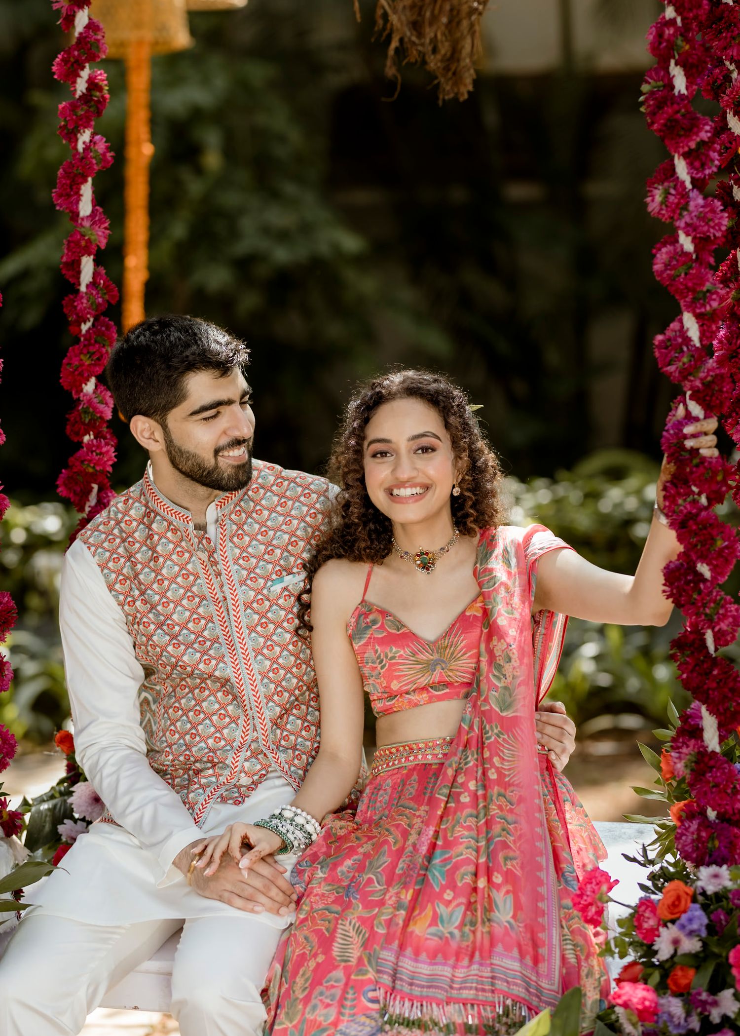 Couple in traditional Indian attire sitting on floral swing, man in embroidered kurta, woman in pink lehenga.