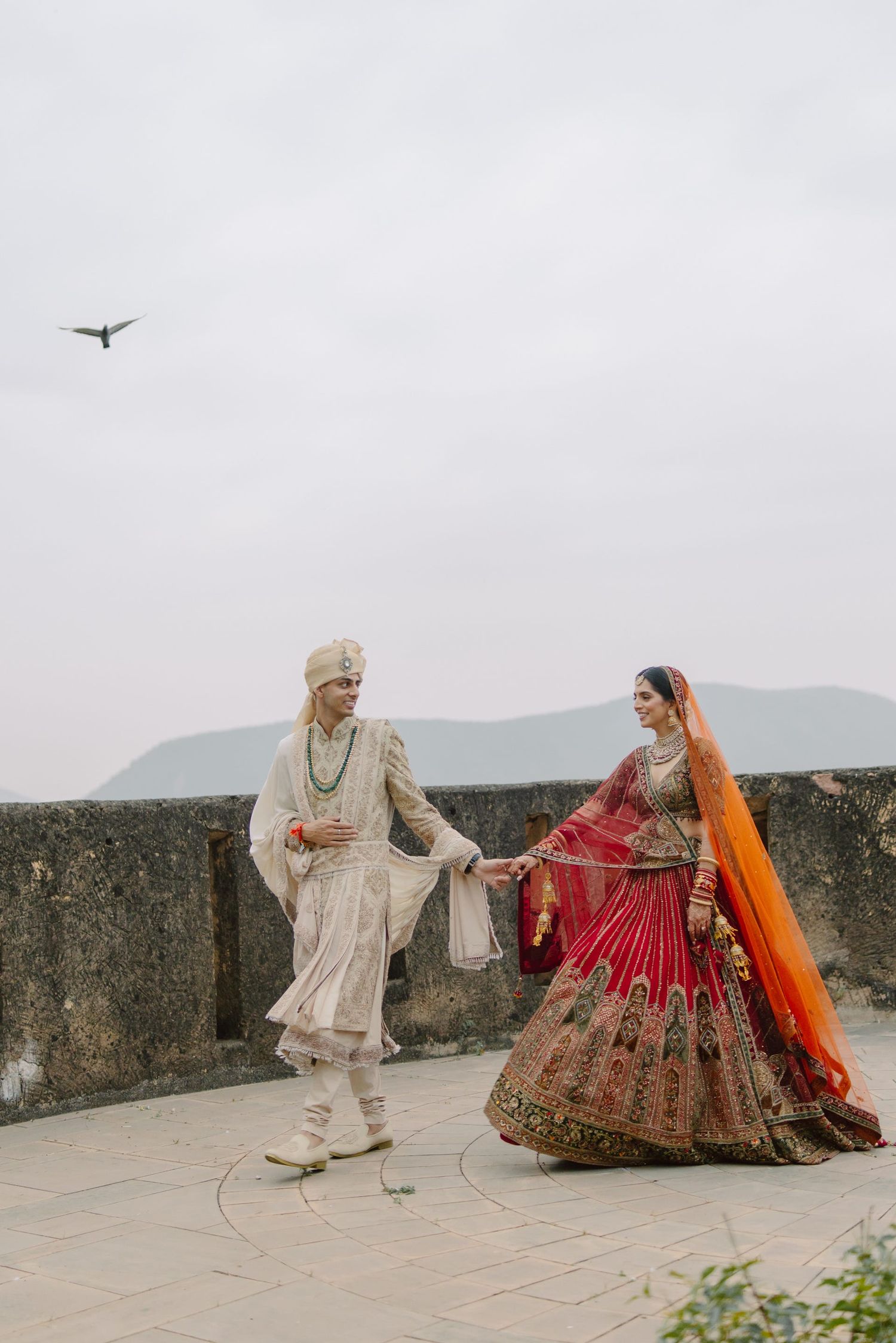 A couple in traditional Indian wedding attire dances on a terrace with mountains and airplane in background.