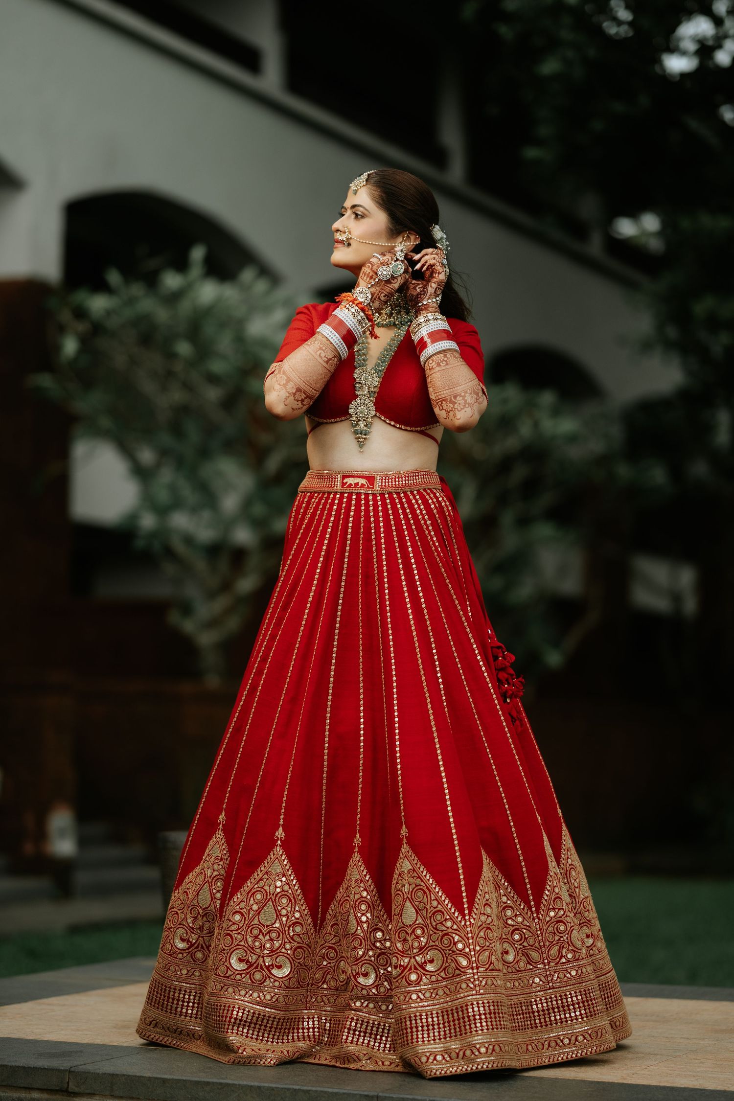 A bride in a stunning red embroidered lehenga with gold detailing adjusts her jewelry outdoors.