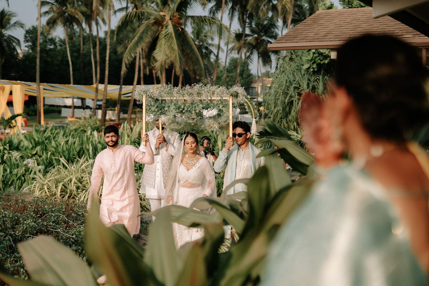 Wedding procession in lush tropical garden with guests in pastel outfits celebrating an Indian ceremony.