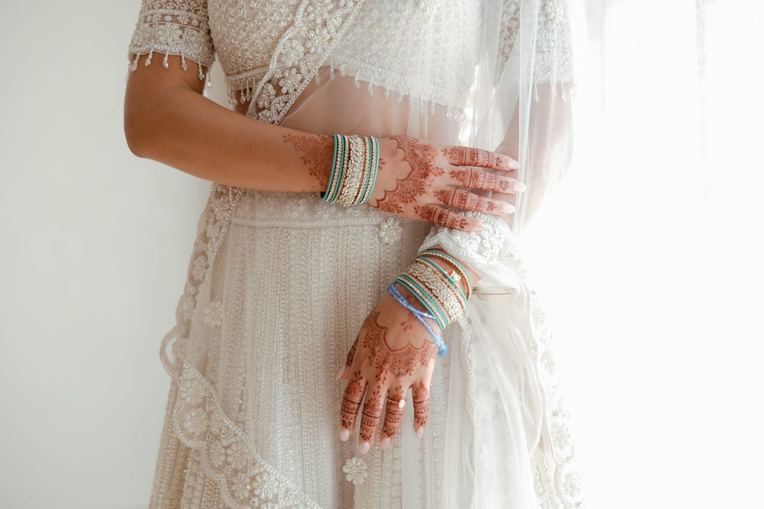Bride in white lace dress with colorful bangles and henna mehndi on hands, holding a water bottle.