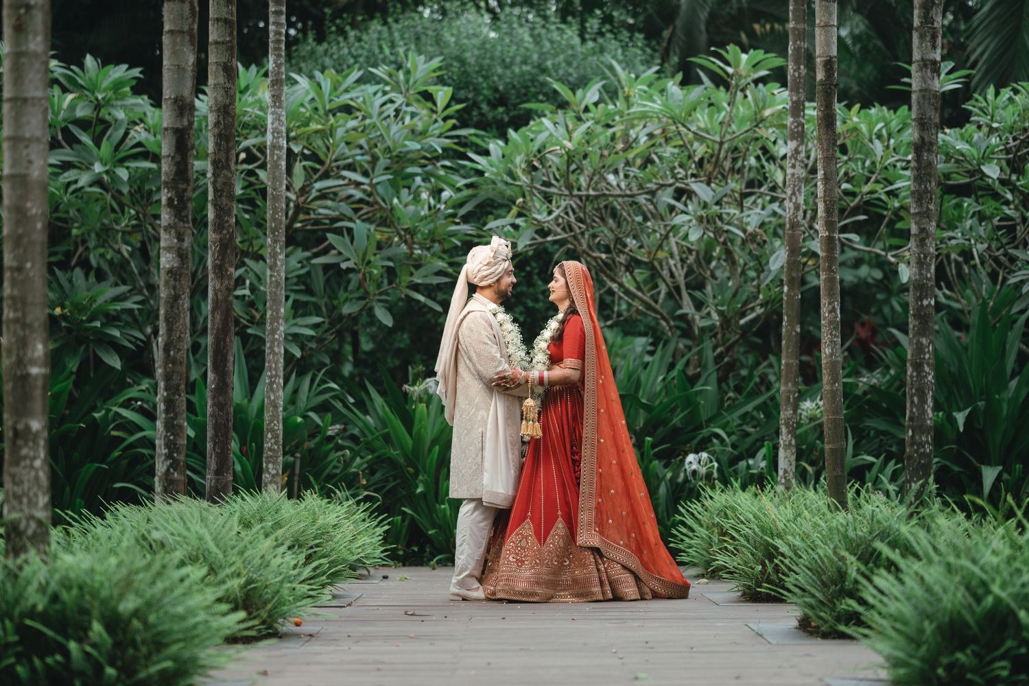 Indian bride in red lehenga and groom in cream sherwani stand together in lush garden during wedding ceremony.