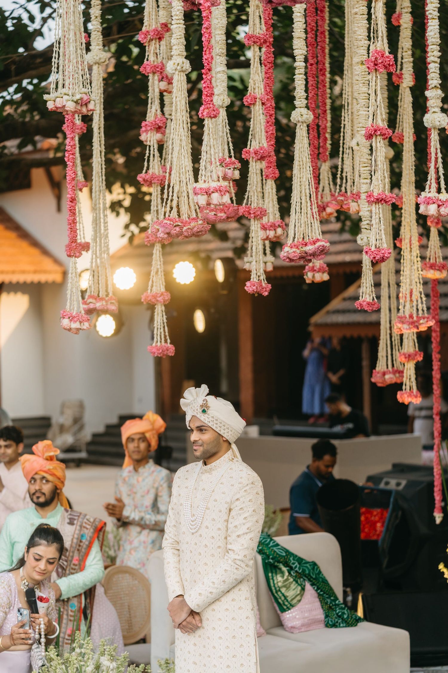 Groom in white sherwani stands under floral chandelier with pink roses at an Indian wedding ceremony.