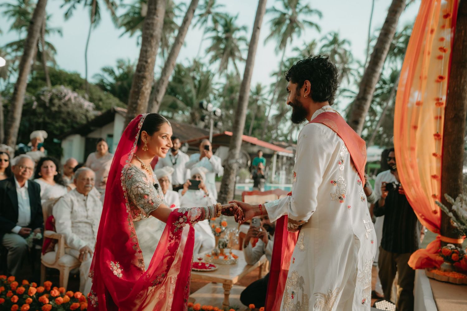 Bride and groom exchange vows at an outdoor Indian wedding ceremony with tropical palm trees and guests.