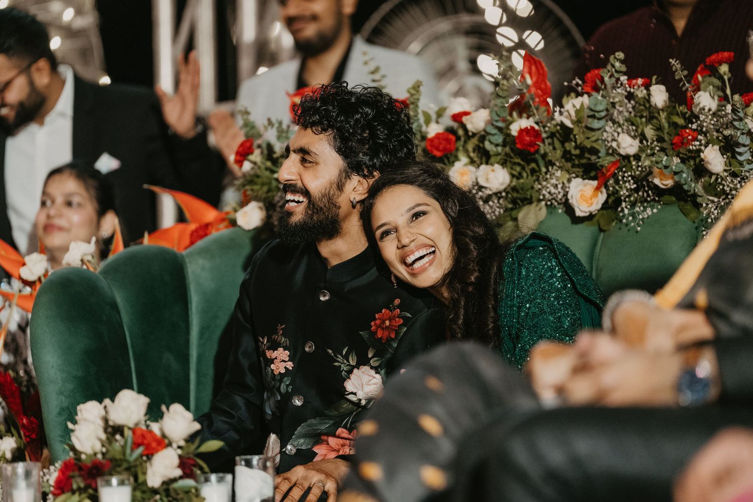 Couple laughing joyfully at a wedding reception surrounded by red and white floral arrangements.