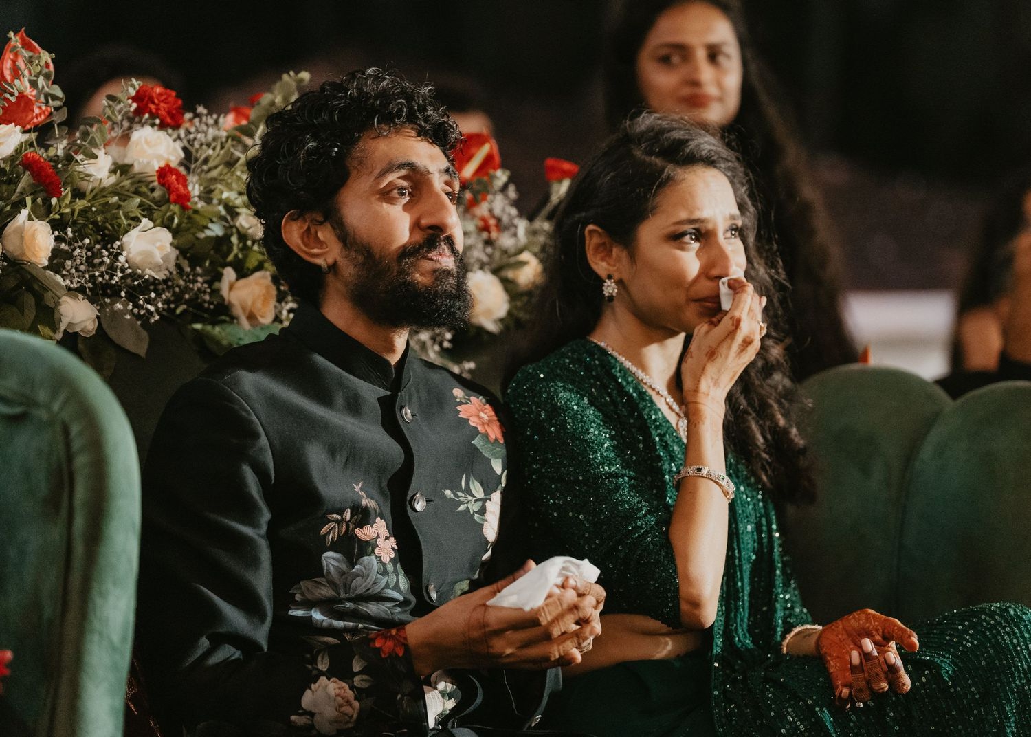 Couple in elegant Indian attire sitting together at a festive event, woman in green sequin saree, man in black embroidered suit.