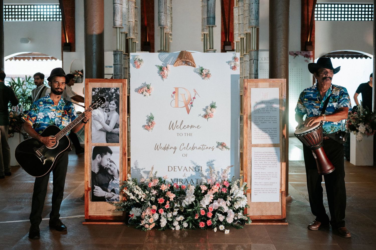 Two musicians in floral shirts perform beside a floral wedding welcome sign for Devang and Priya's celebration.