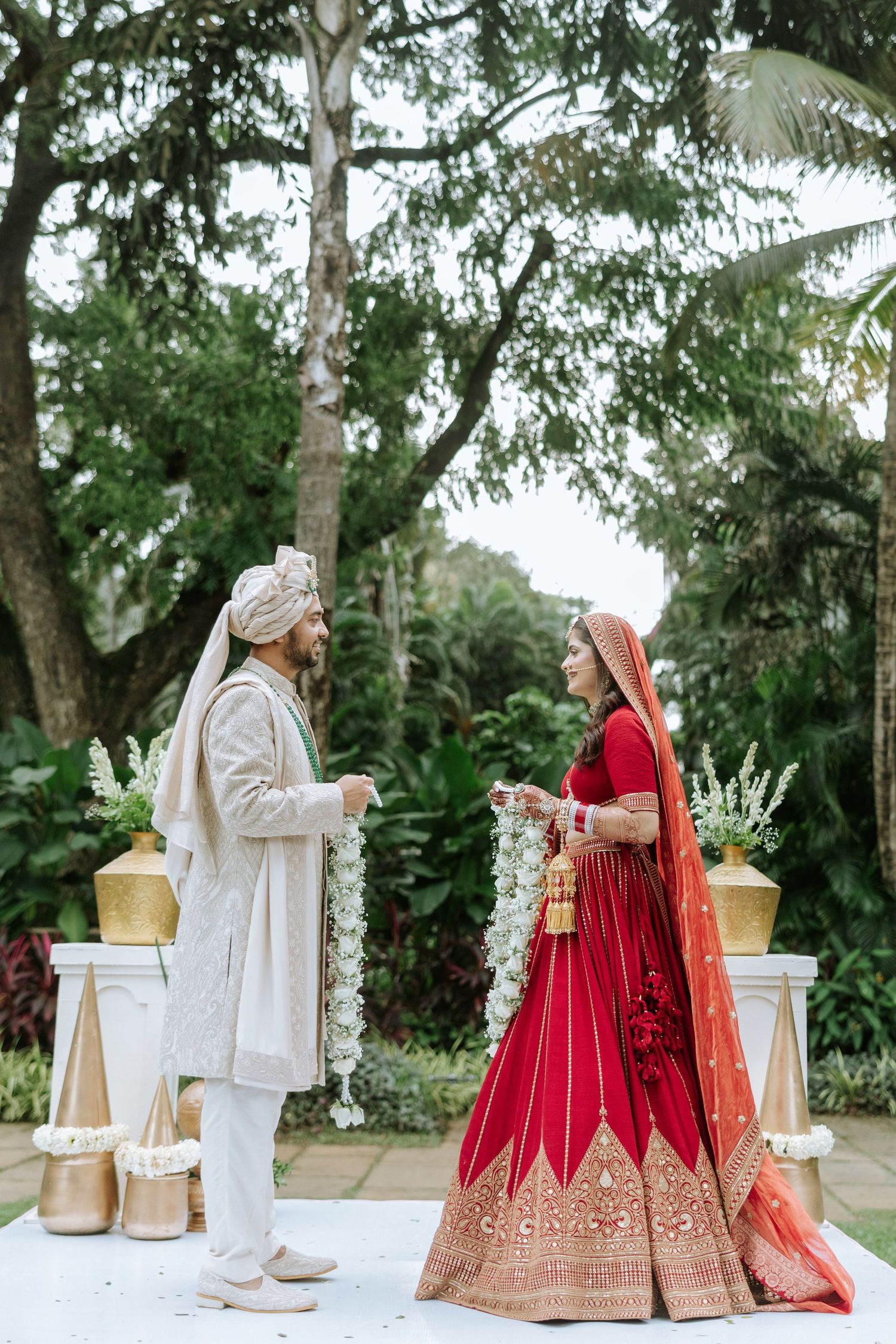 Indian bride in red lehenga and groom in white sherwani exchange garlands at outdoor Hindu wedding ceremony.