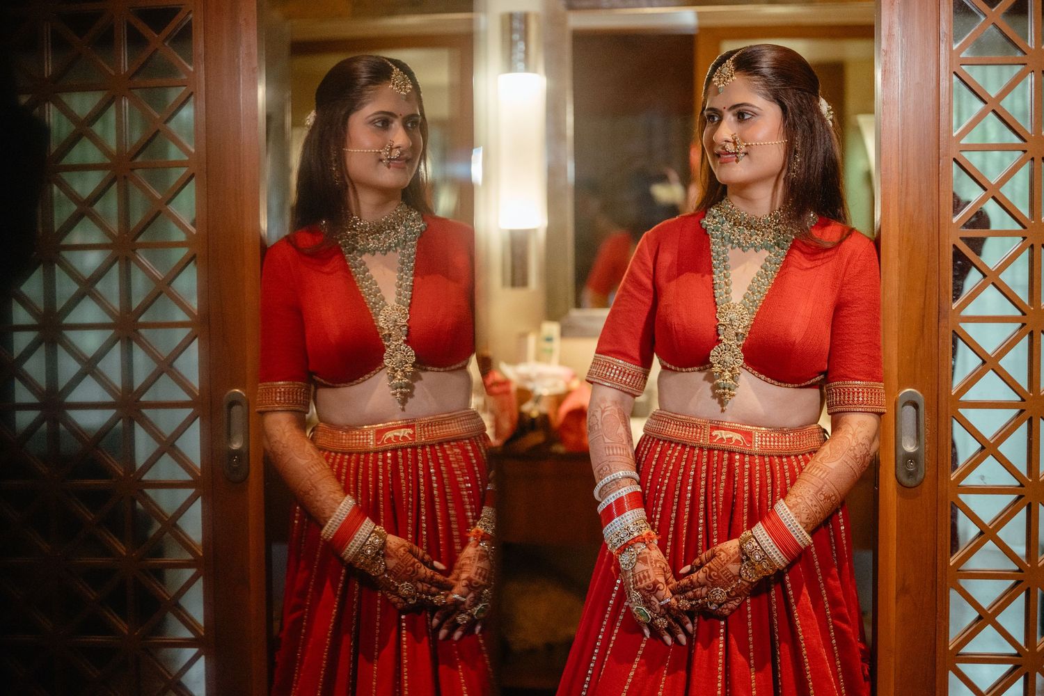 Indian bride in red lehenga with gold jewelry poses before mirror showing stunning mehndi hands.