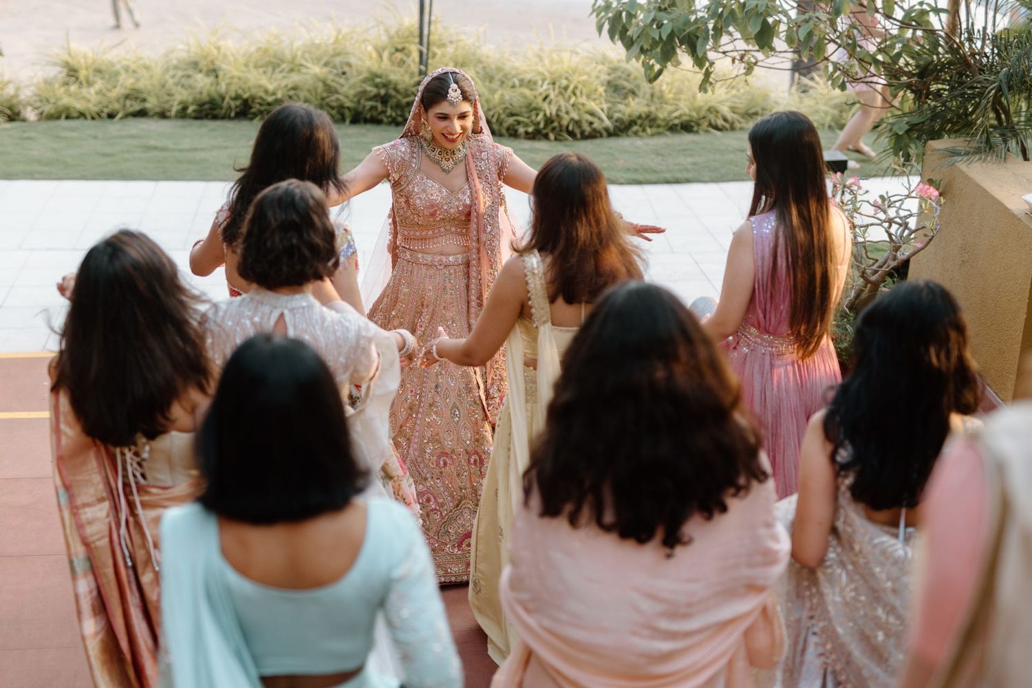 A group in pastel and blush-colored formal attire gather outdoors near a waterfront setting.