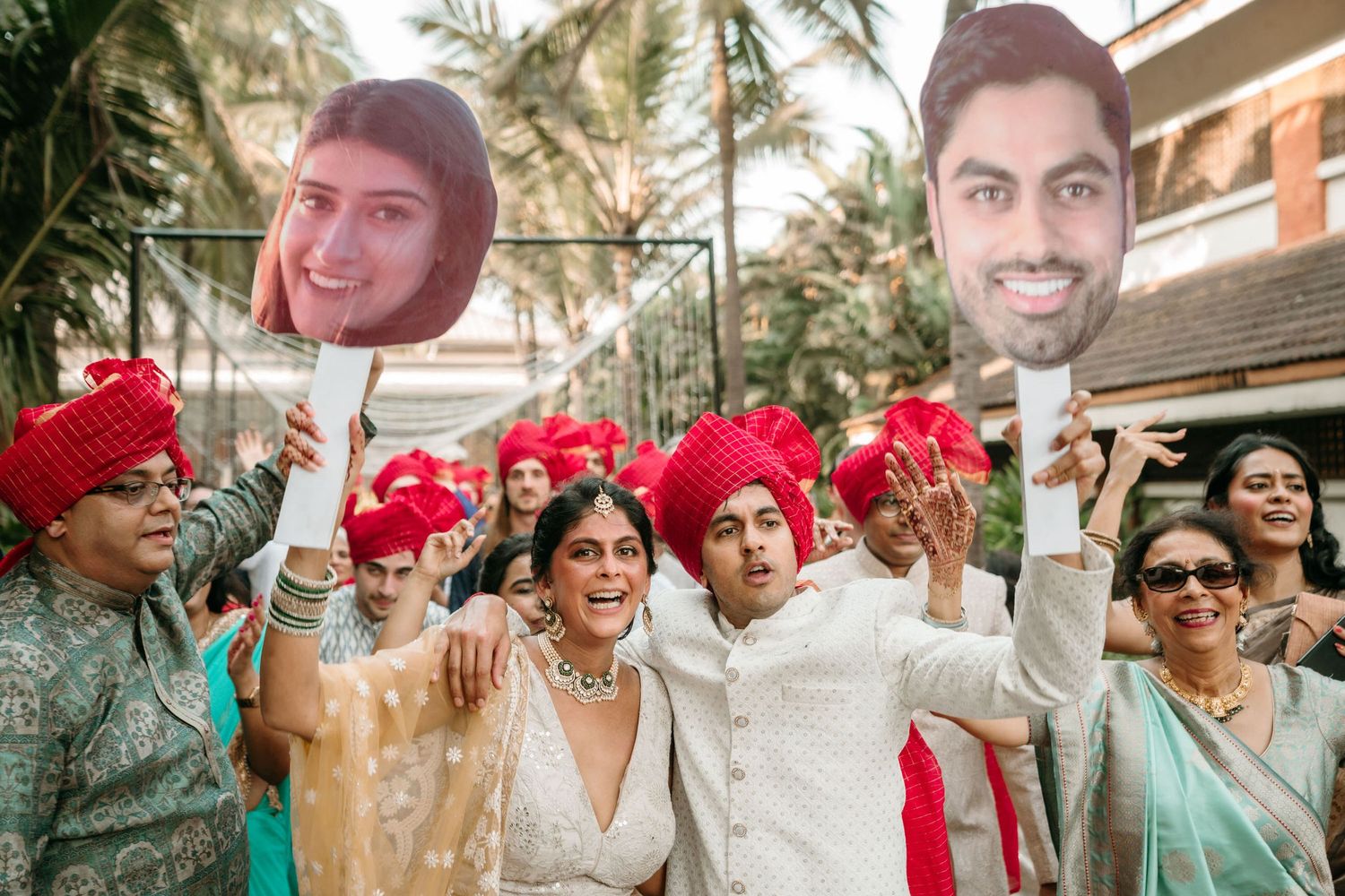 Wedding guests in traditional Indian attire holding photos on sticks during a celebration with palm trees in background.