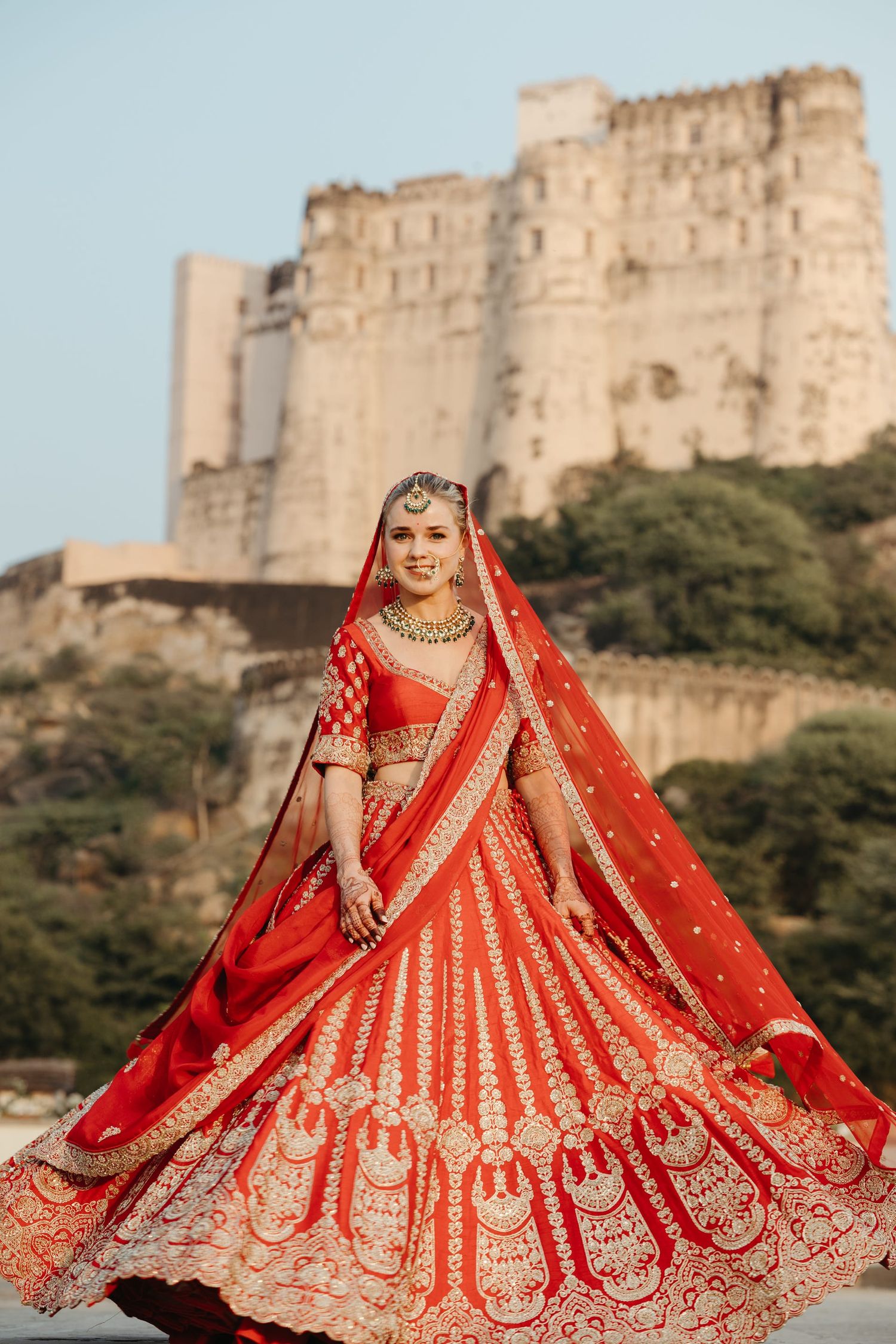 Stunning red and gold bridal lehenga twirling against historic fort backdrop in traditional Indian wedding photoshoot.