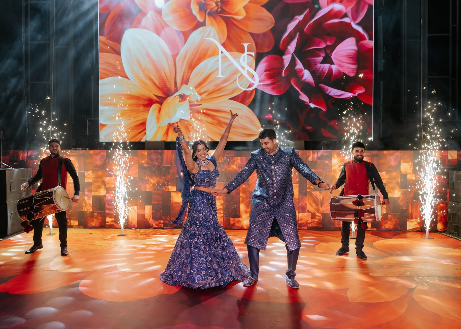 Bride and groom perform first dance on stage with pyrotechnics and floral backdrop at Indian wedding reception.