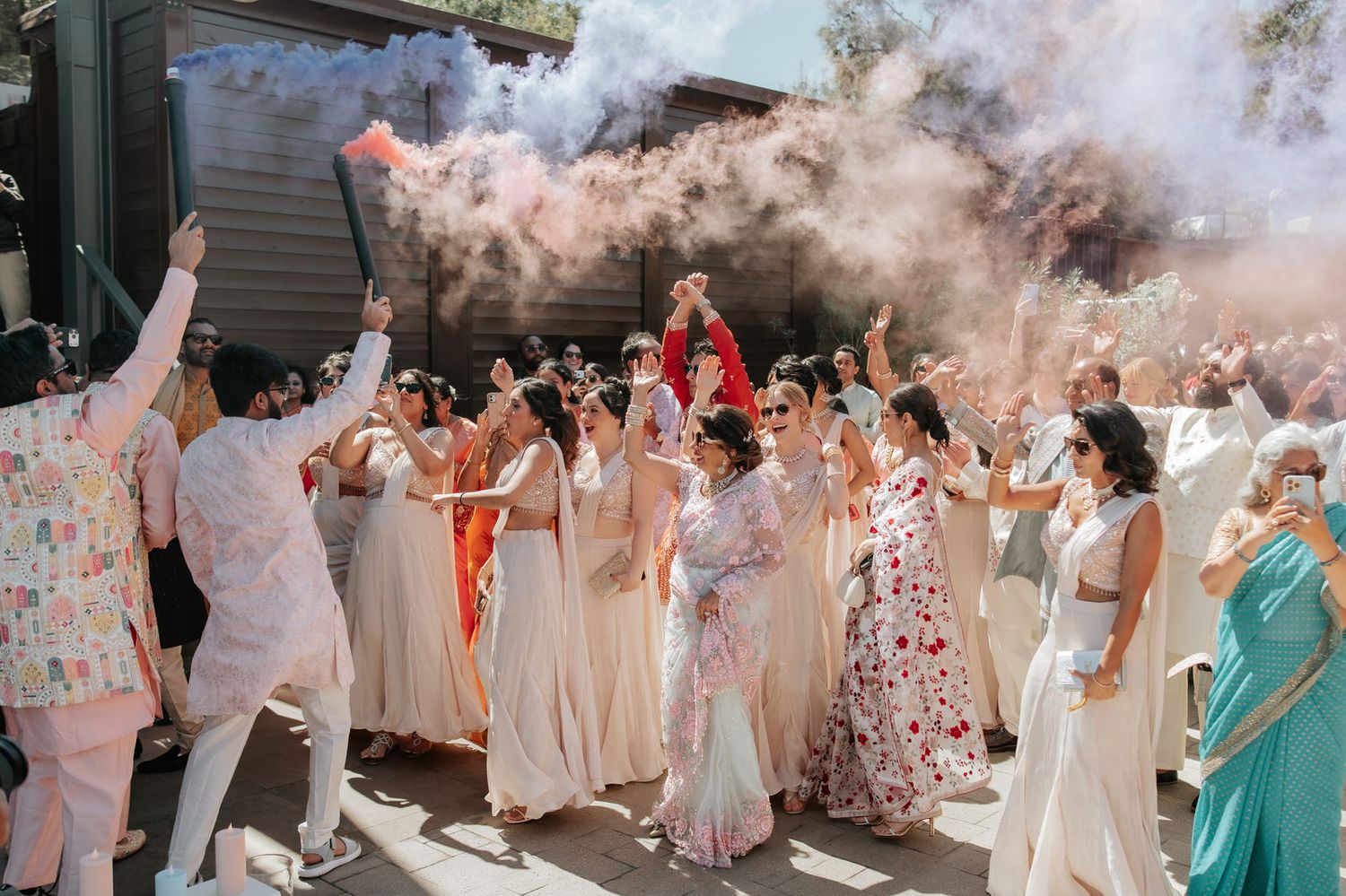 Colorful smoke bombs fill the air as wedding guests in white and pastel sarees celebrate outdoors in a joyful procession.