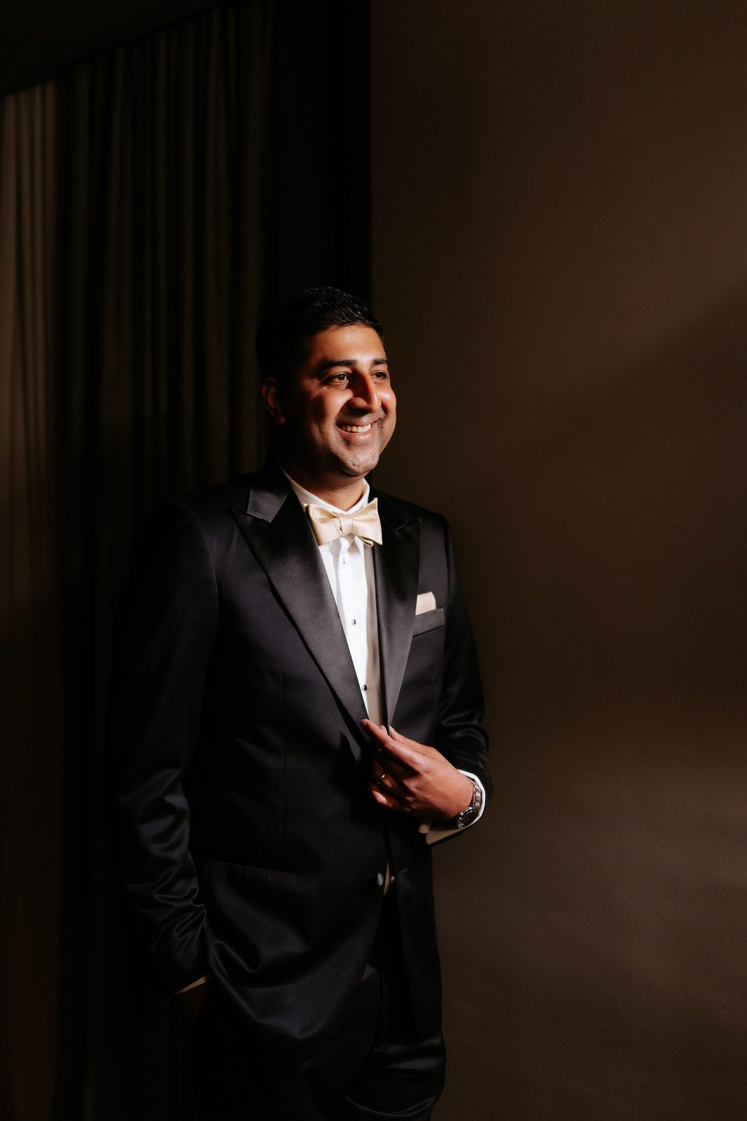 Handsome Indian groom smiling confidently in black tuxedo with bow tie against dark dramatic background.