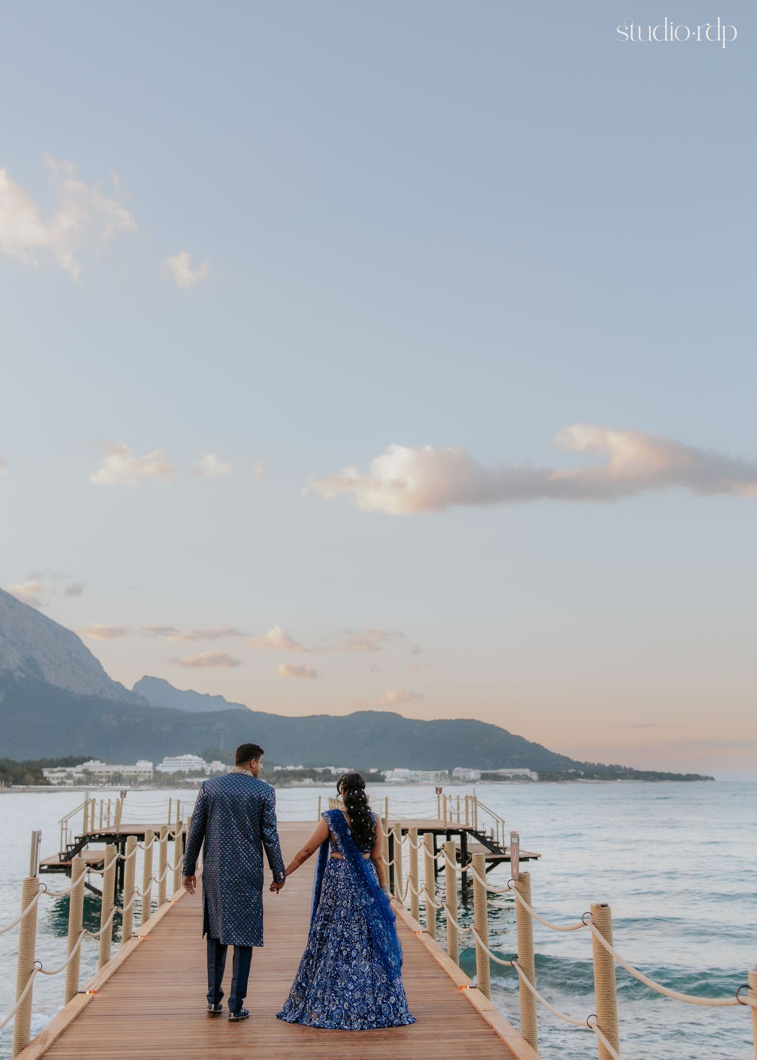 Couple in blue Indian attire holding hands on a wooden pier overlooking the ocean at sunset.