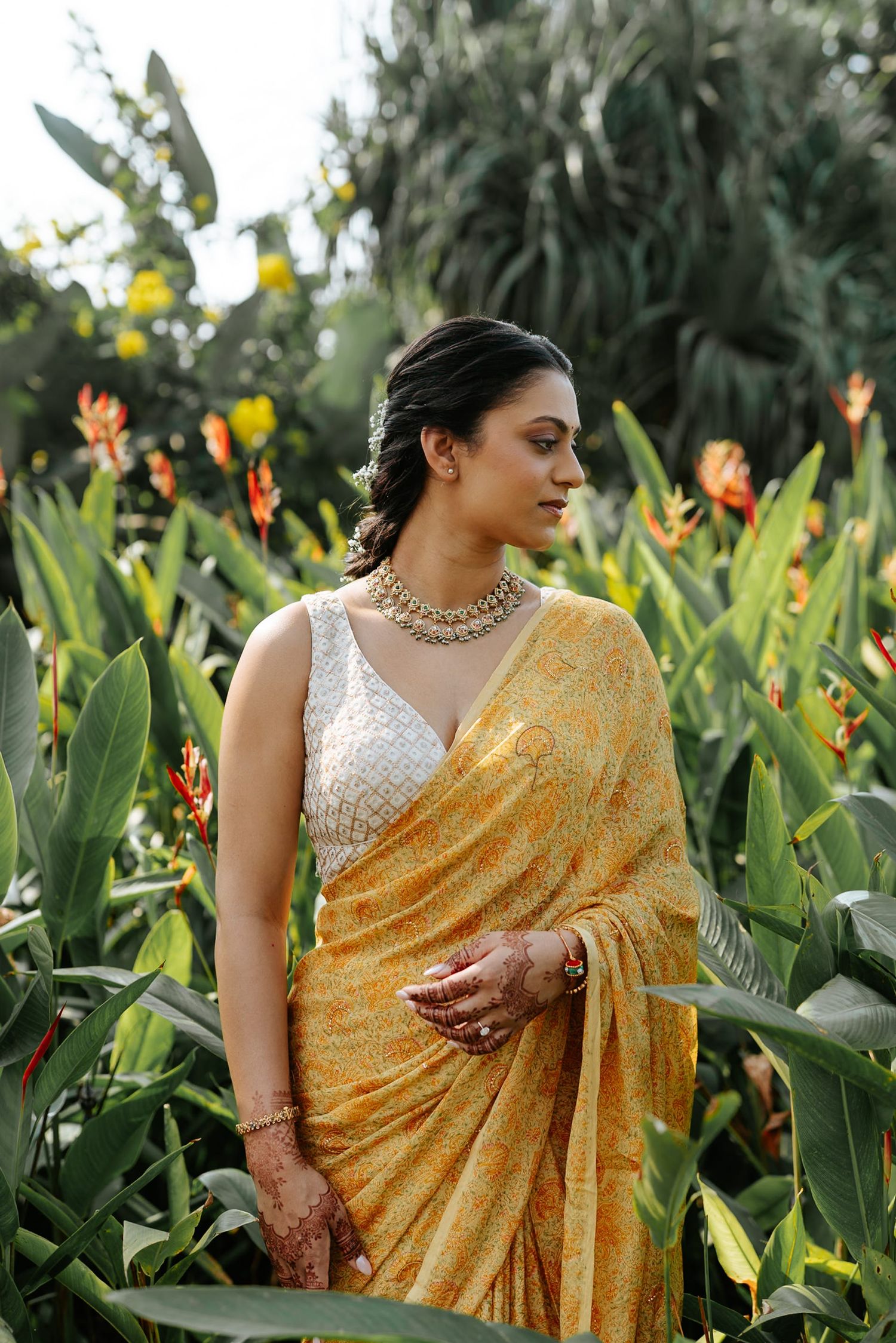 Woman in yellow floral saree and gold jewelry standing among tropical flowers in a lush garden.