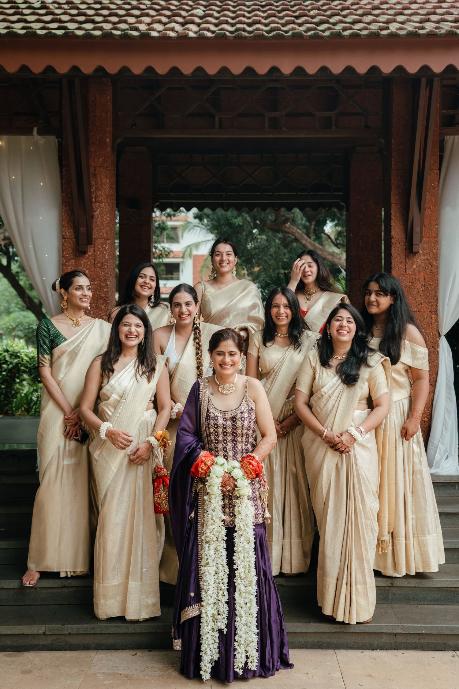 Bride in purple saree holds floral garland surrounded by bridesmaids in cream sarees at traditional Indian wedding.