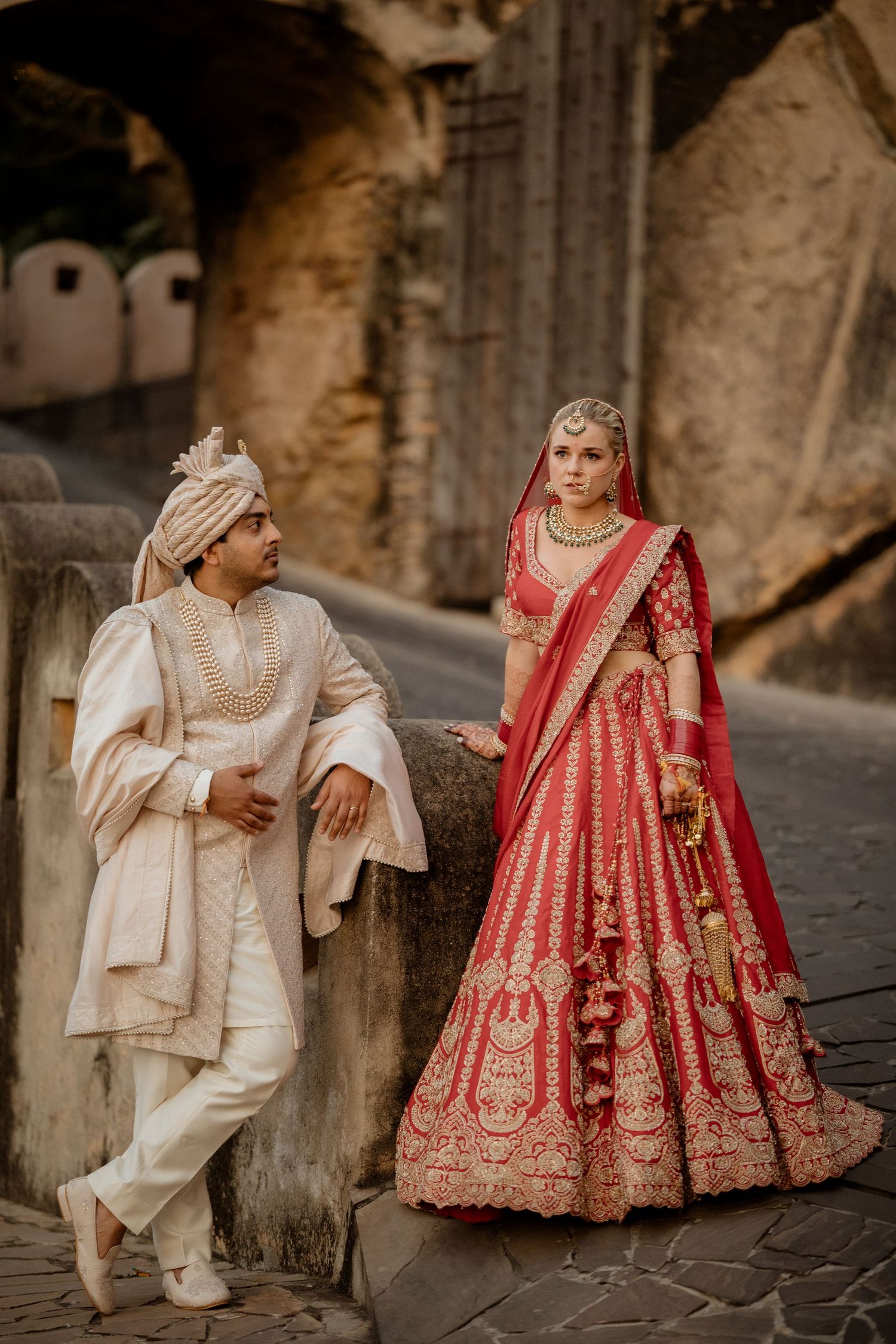 Wedding couple in traditional Indian attire posing against stone architecture.