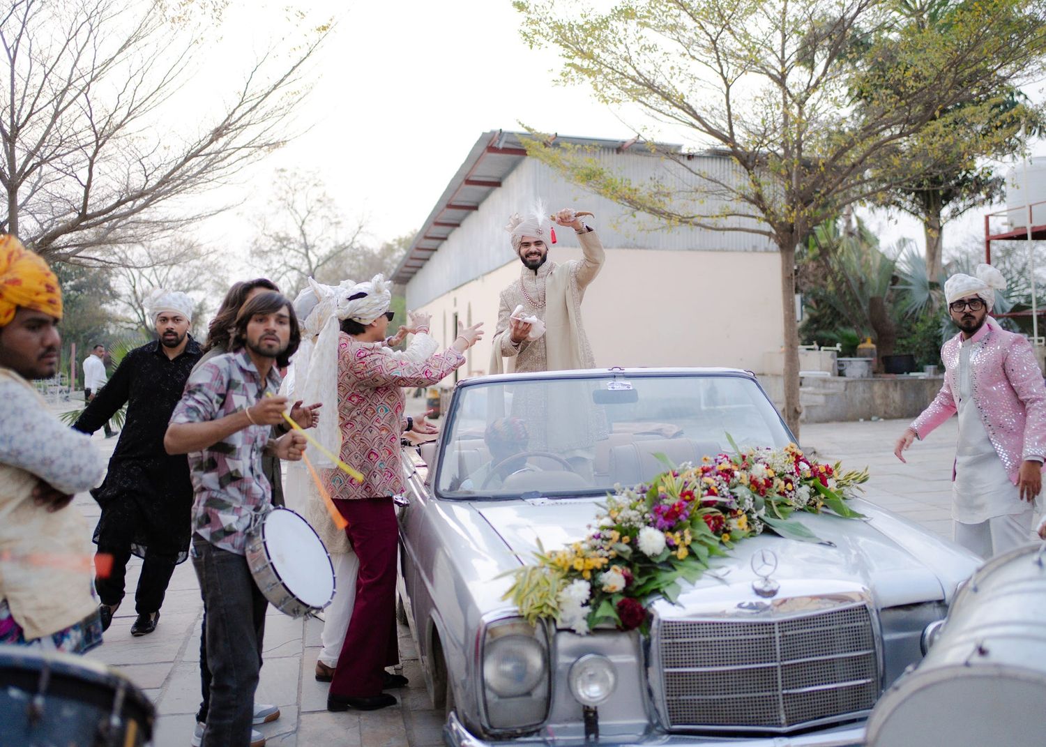 Groom stands on vintage Mercedes-Benz at Indian wedding baraat celebration with drummers and guests in traditional attire.